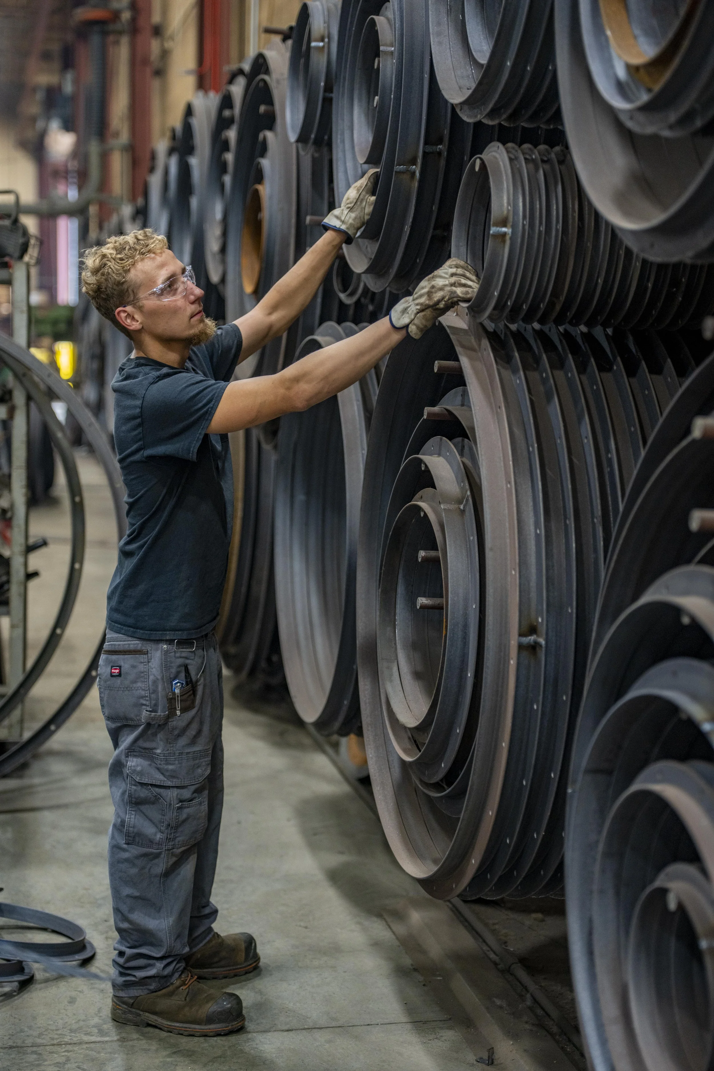 A worker inspecting large metal machine parts on shelves in an industrial factory or workshop.