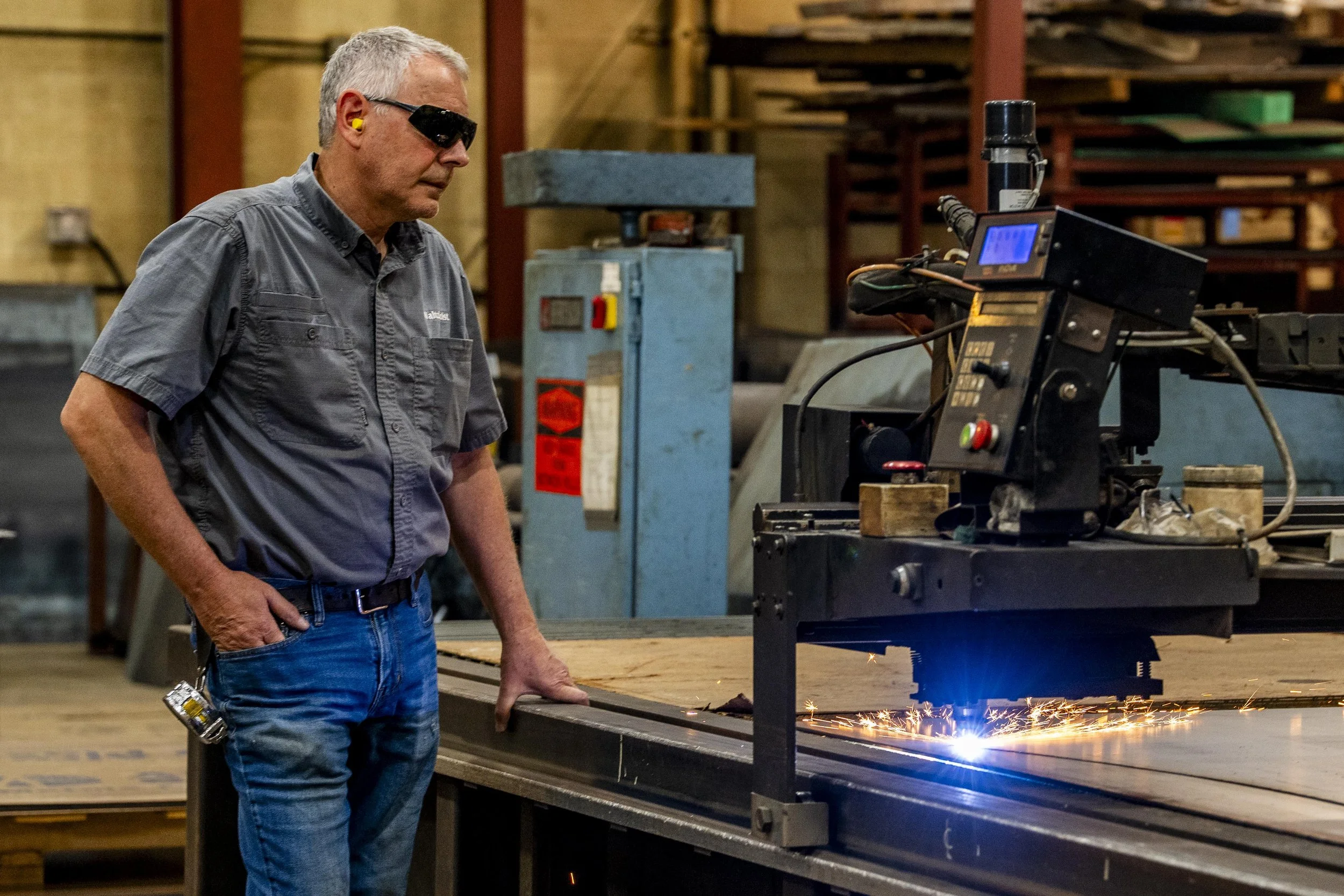 A man wearing safety glasses and a gray work shirt stands next to a metal cutting machine in a workshop, watching sparks fly from the cutting process.