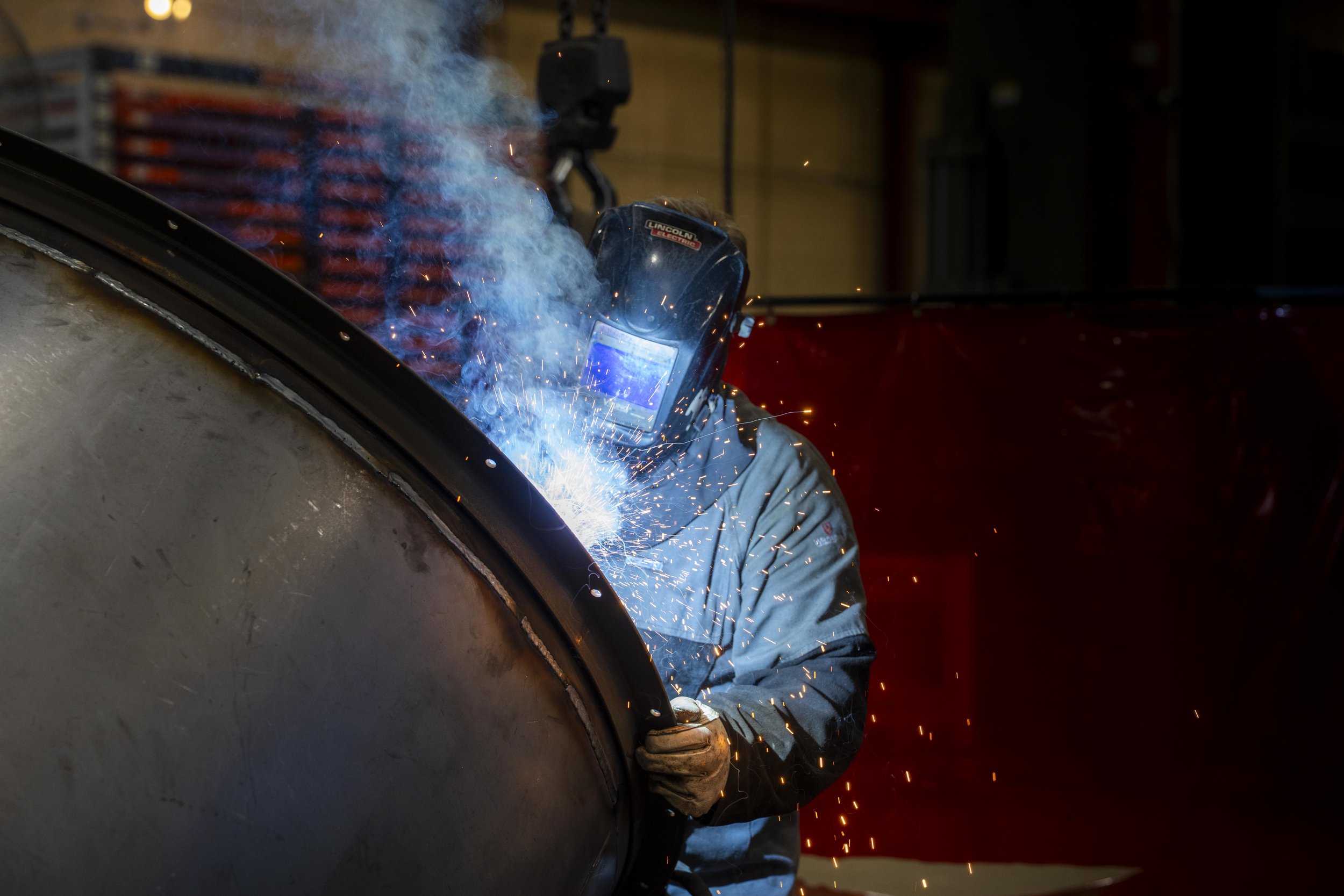 A person wearing a welding helmet and gloves welding a large metal pipe, with sparks and blue welding light emitting from the process, in an industrial workshop.