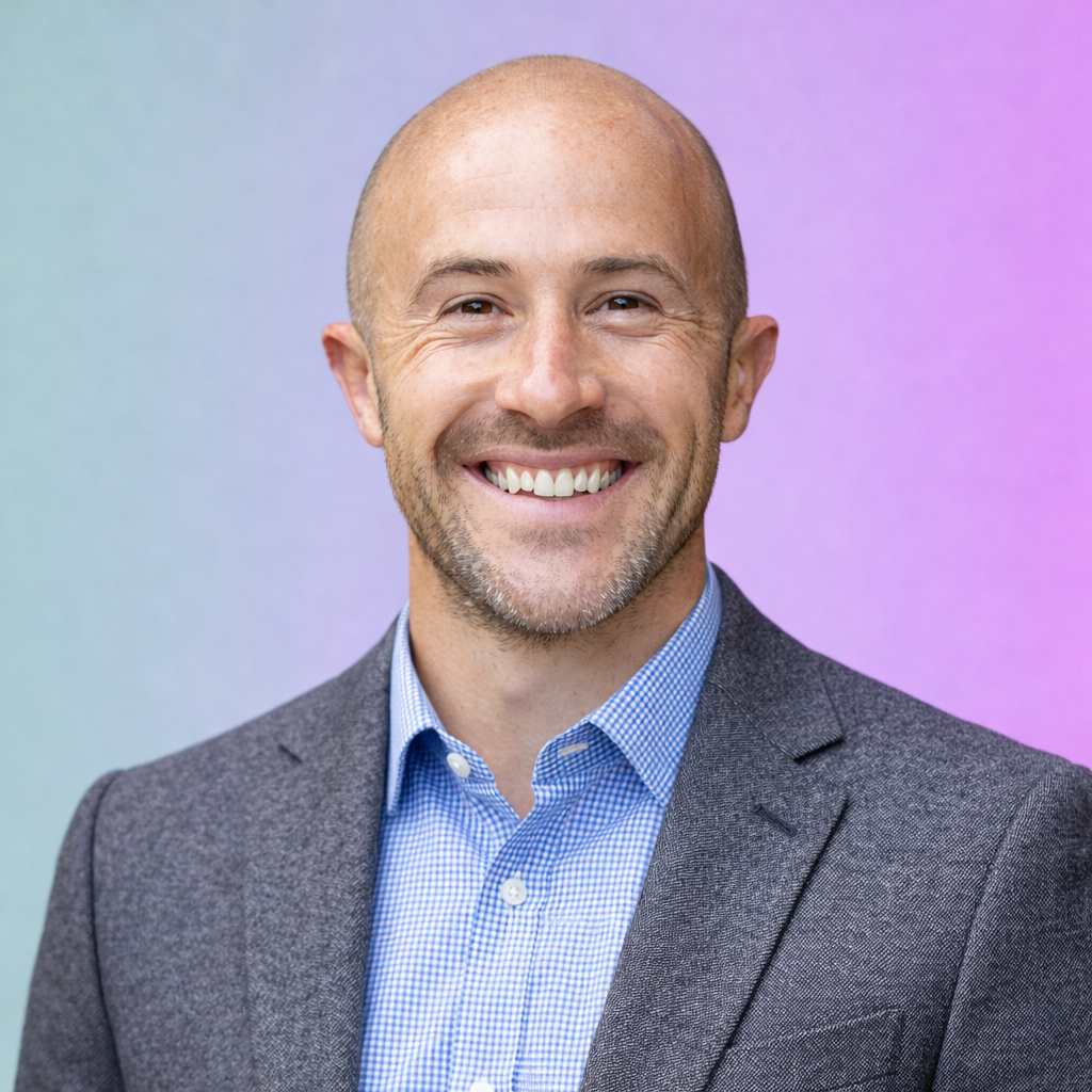 Close-up of smiling man with a shaved head and a beard, wearing a maroon shirt, outdoors with green foliage in the background.