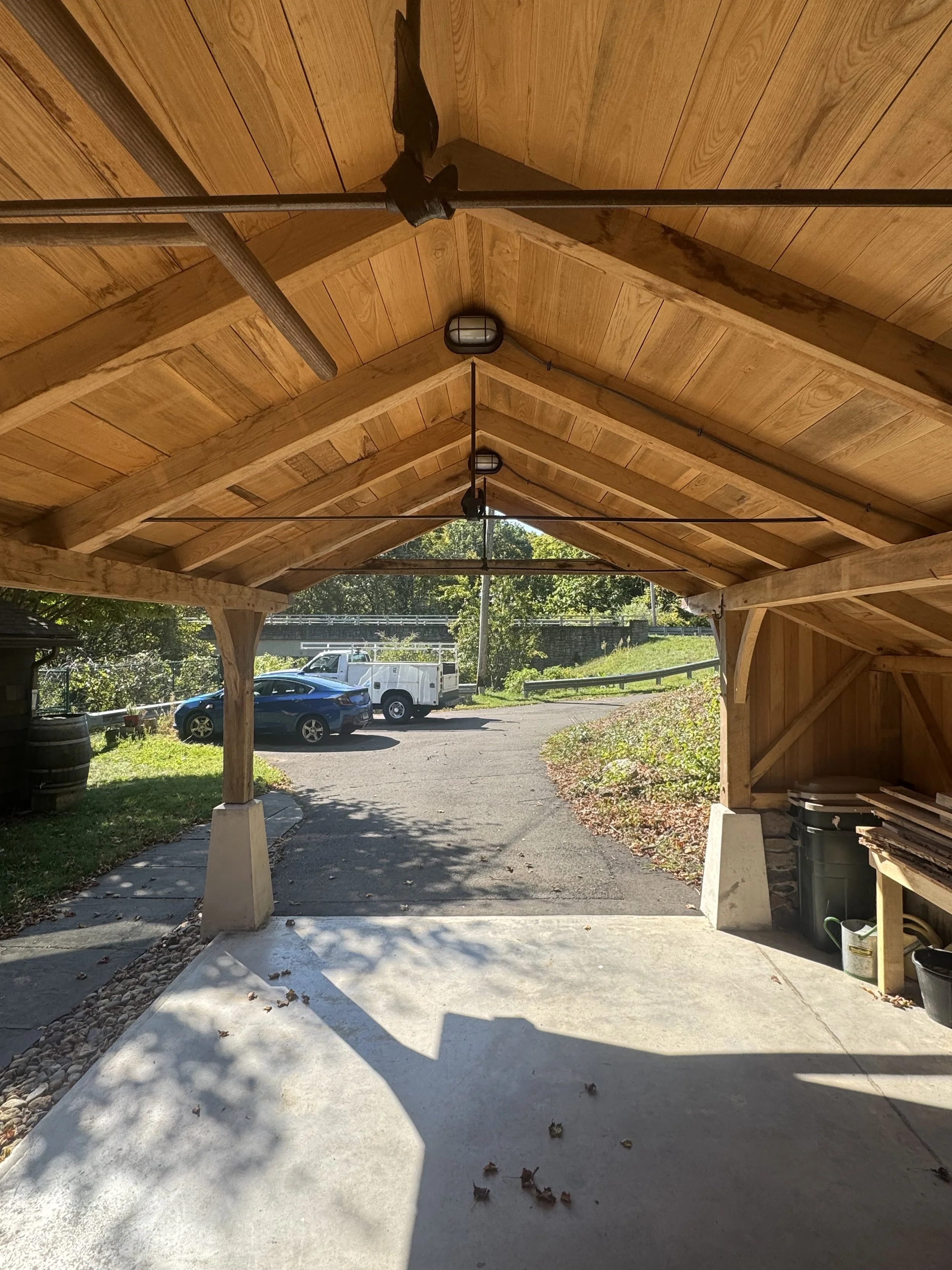 Under a wooden carport with a wooden ceiling and support beams, view of parked cars and a driveway leading to a grassy area and trees.