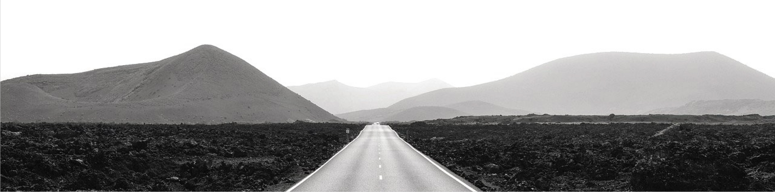 Black and white photo of a straight road extending into the distance through a barren landscape with valley and mountains in the background.