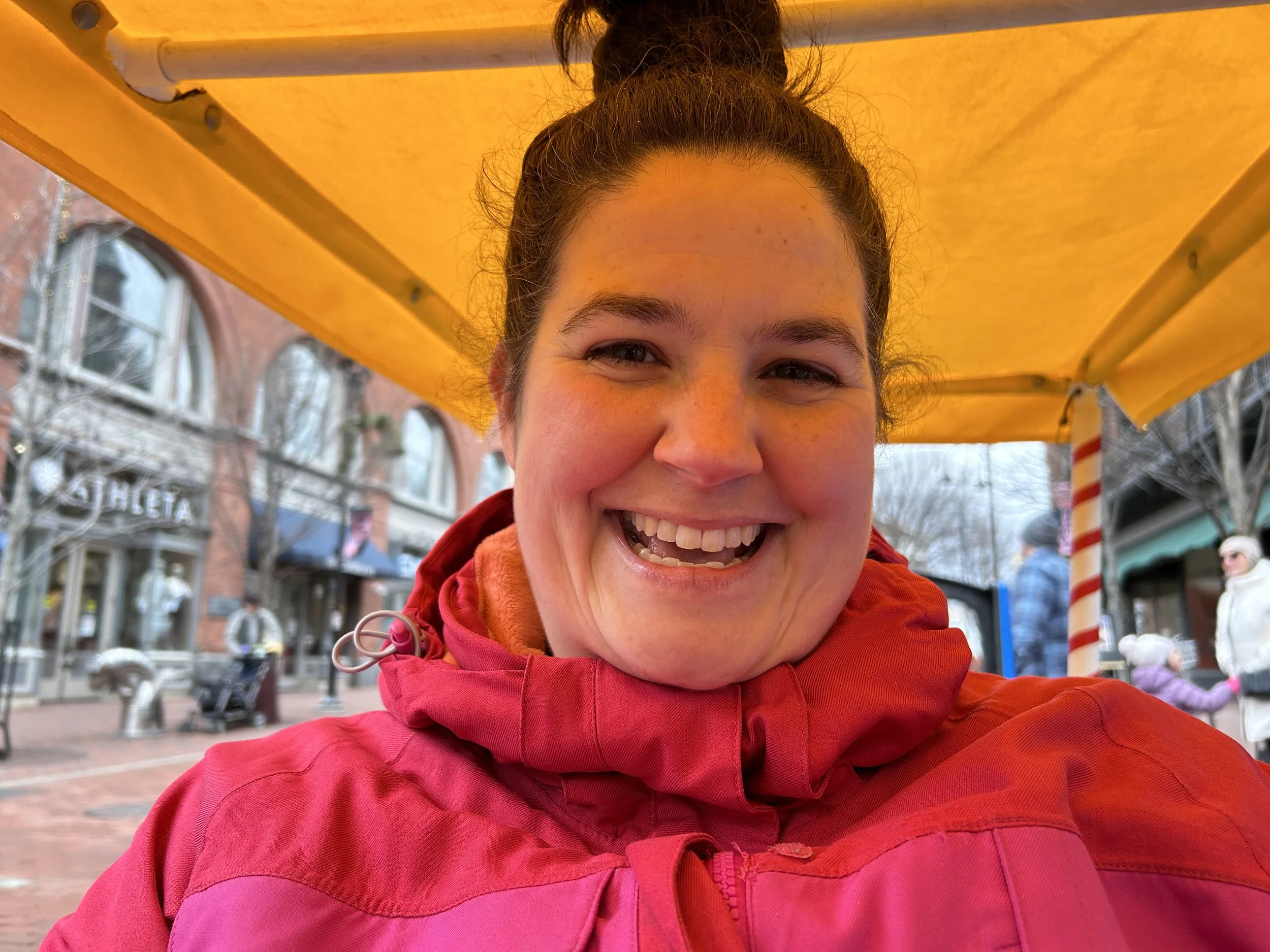 A smiling woman with dark hair in a bun, wearing a red jacket, is taking a selfie under a yellow canopy during winter in an urban area with people and brick buildings in the background.