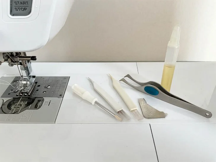 Sewing machine with various sewing tools and accessories on a white surface, including a needle, fabric clips, a seam gauge, and a bottle of sewing lubricant.