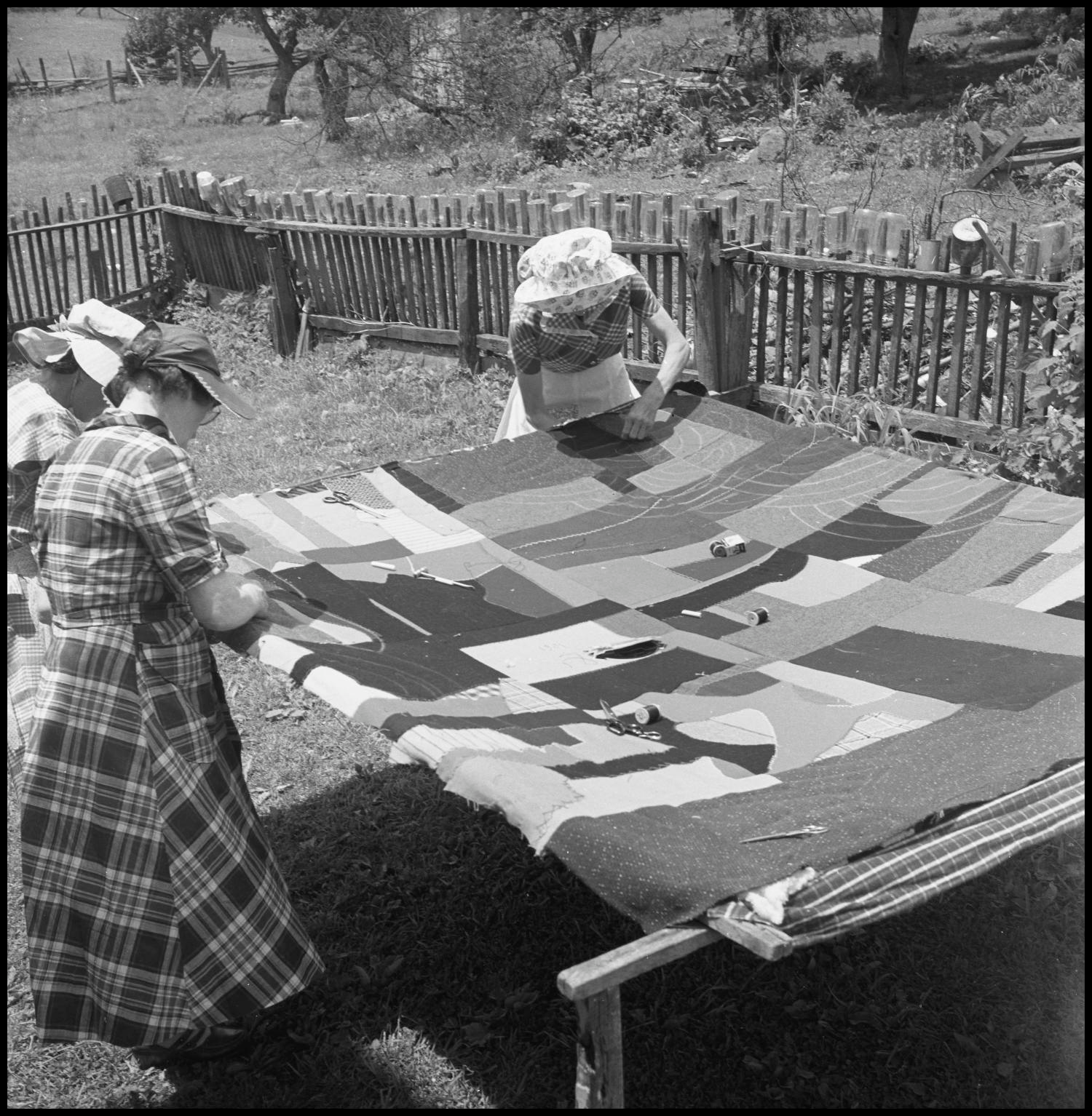 Two women working on quilting outdoors on a large quilt spread over a wooden frame, with a rustic fence and trees in the background.