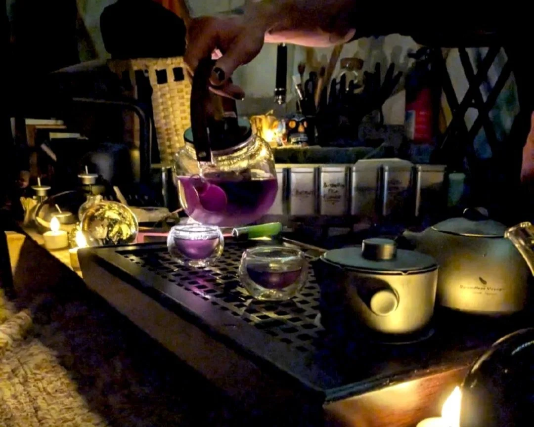 A person making hot purple tea or herbal infusion on a countertop with candles and a teapot, in a cozy, dimly lit kitchen or dining area.