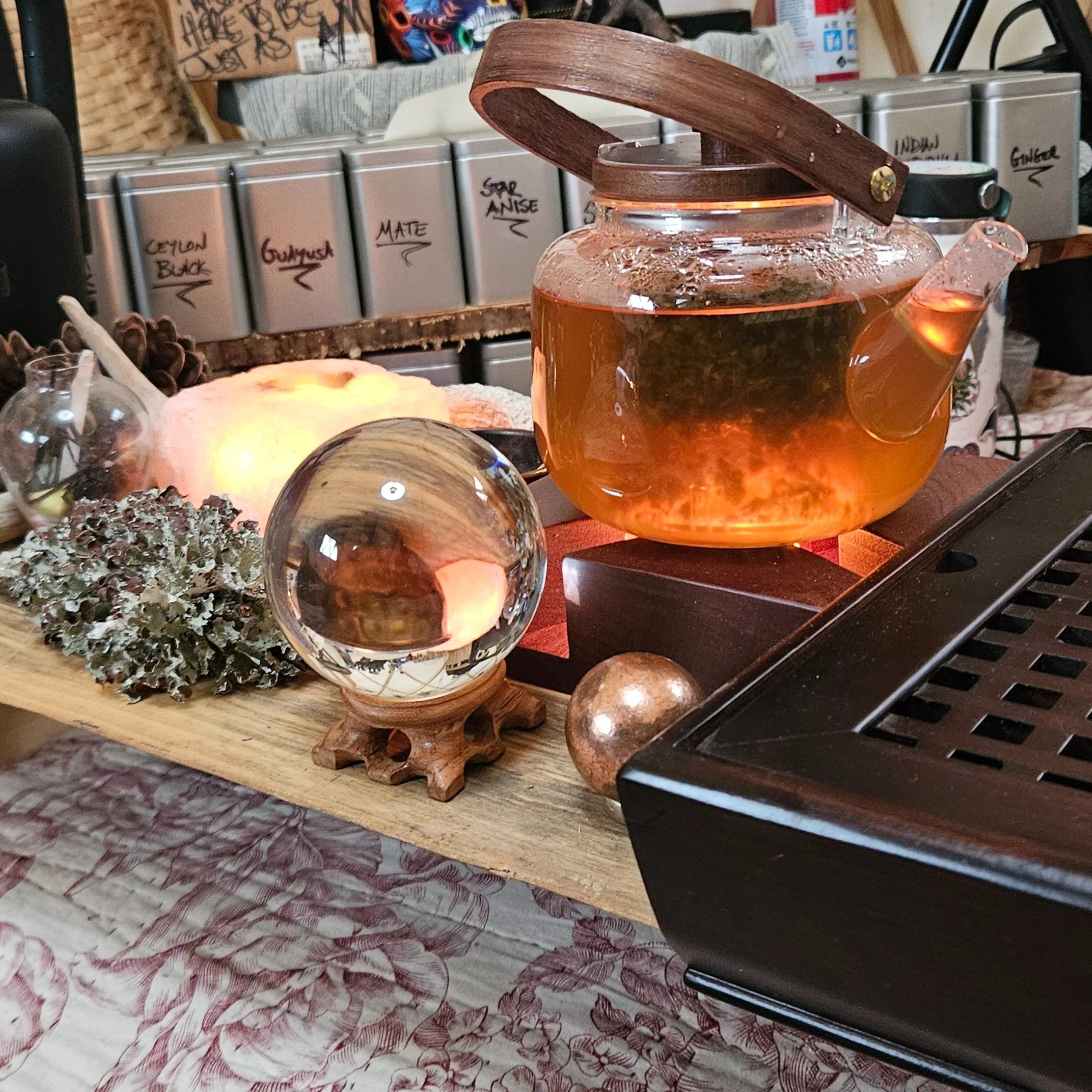 A glass teapot with amber-colored tea on a wooden tray, surrounded by decorative objects including a glass orb, plants, and a Himalayan salt lamp, on a table with a floral tablecloth.