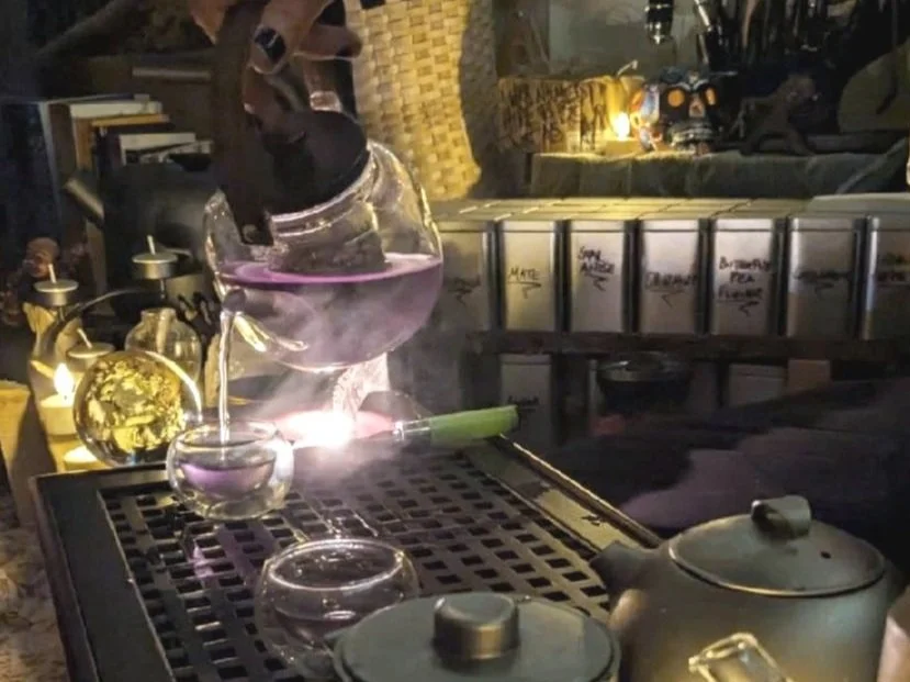 A tea kettle and a glass teapot on a metal table at an outdoor setting during evening, with glassware and decorative items nearby.