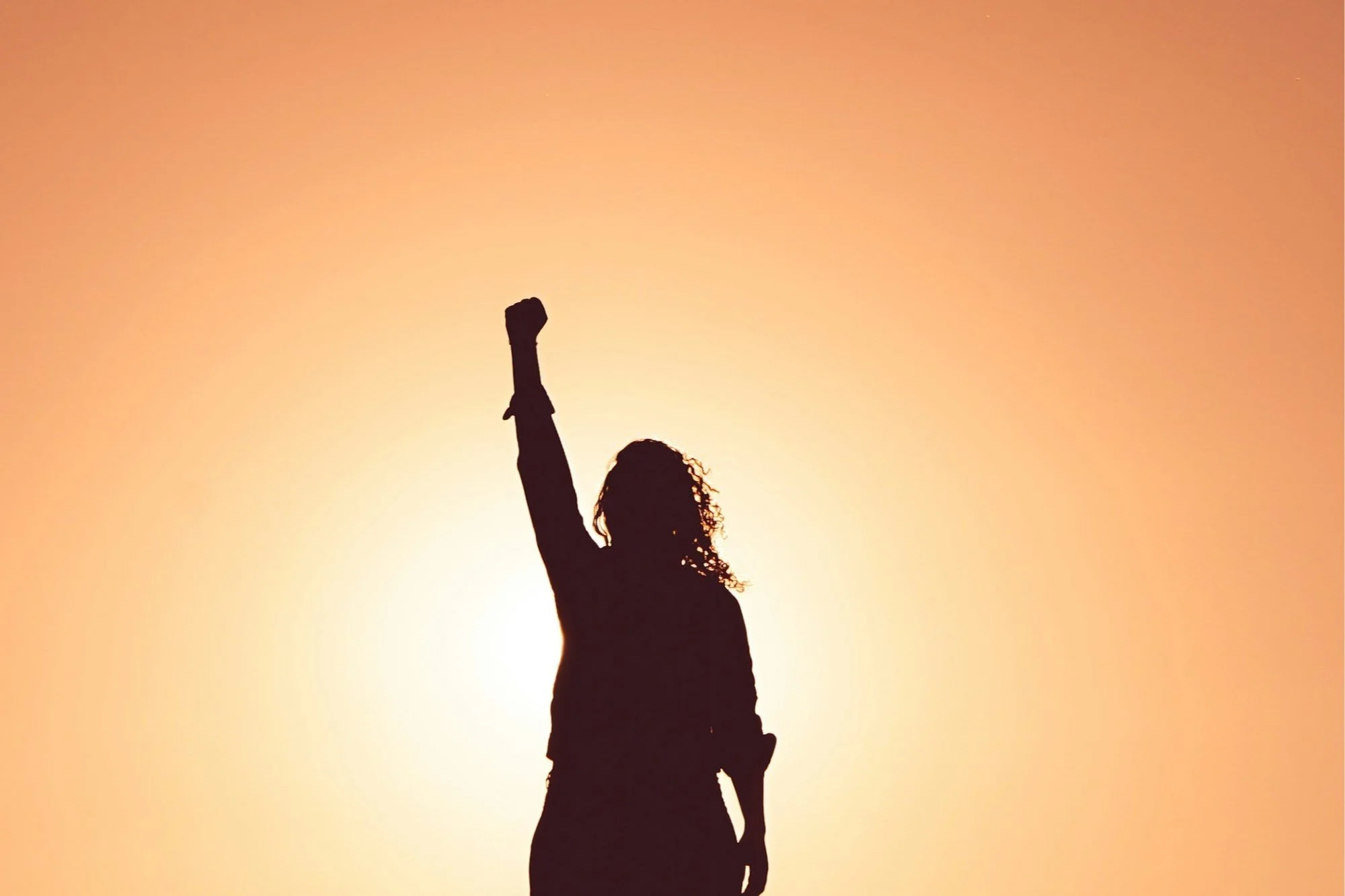 Silhouette of a woman with curly hair raising her right arm in victory against a sunset background.