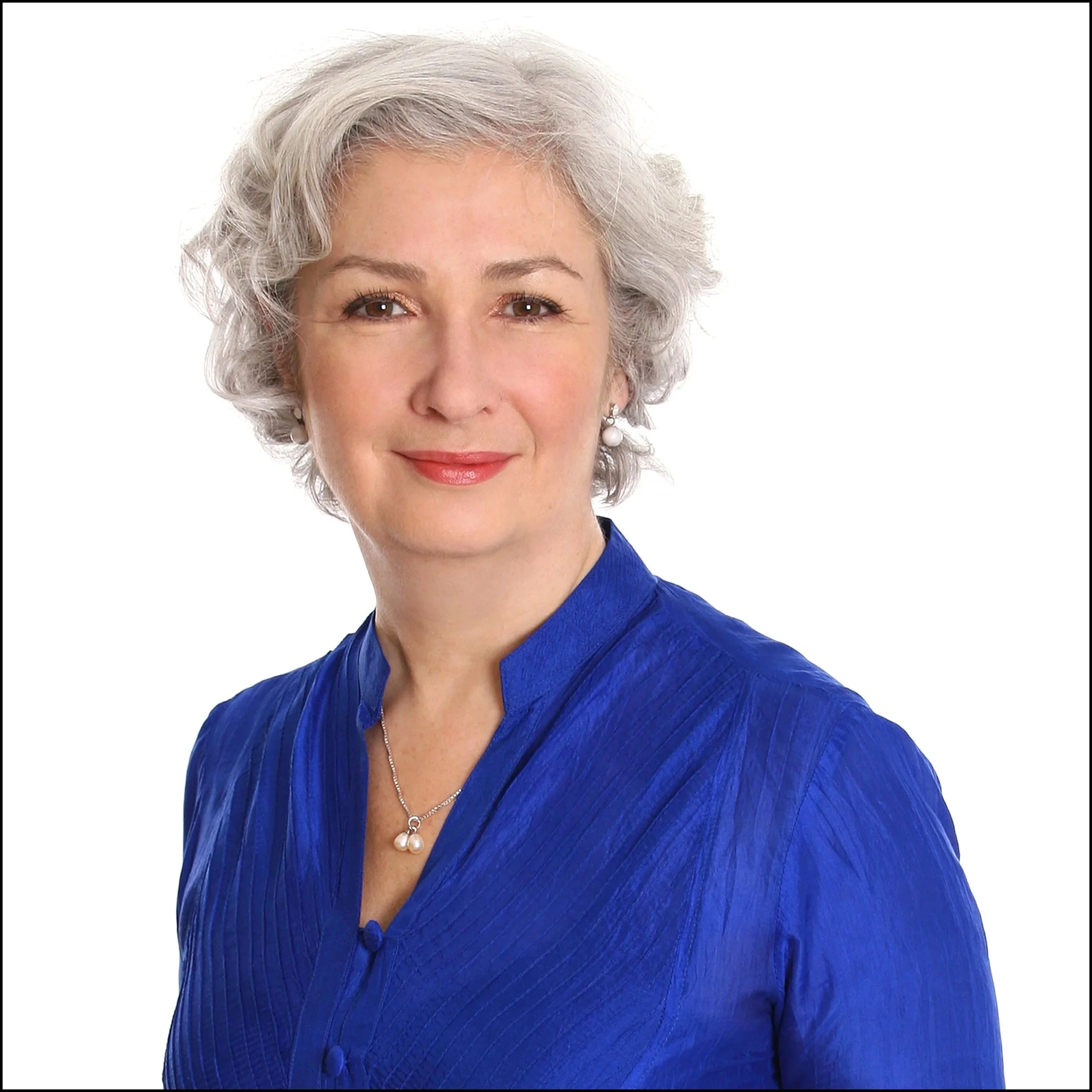 A woman with short, curly gray hair and light skin, smiling, wearing a royal blue blouse with a collar and pearl jewelry, against a white background.