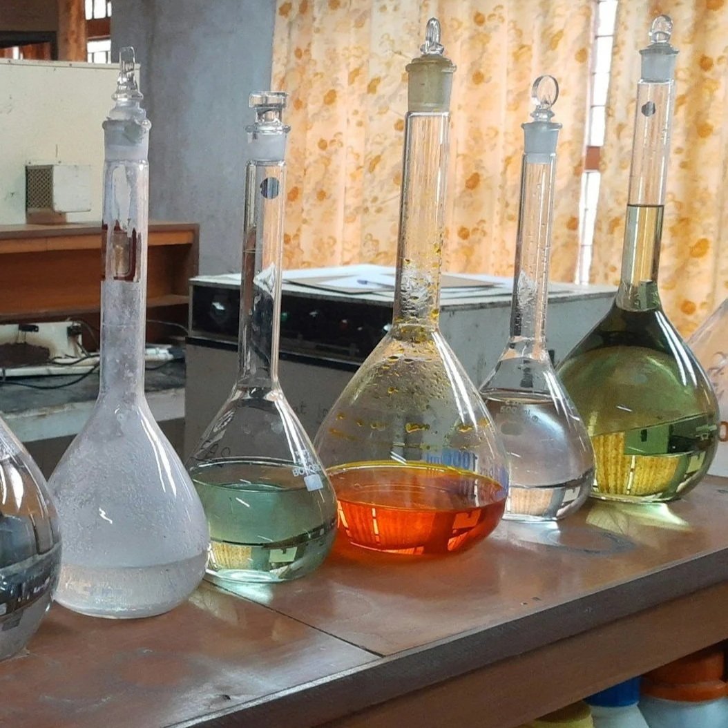 Five glass flasks with various colored liquids on a laboratory table, with a stove and kitchen curtains in the background.