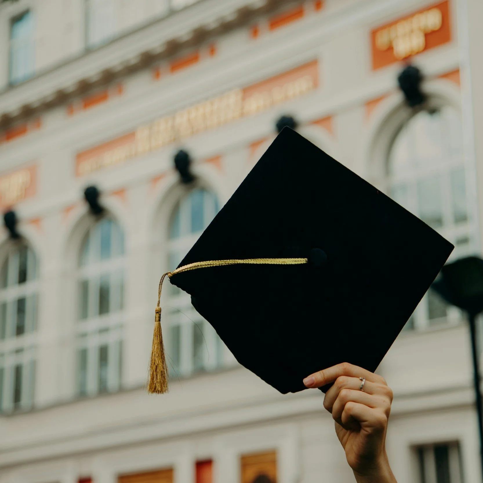 A hand holding up a black graduation cap with a gold tassel in front of a building with white walls and multiple arched windows.