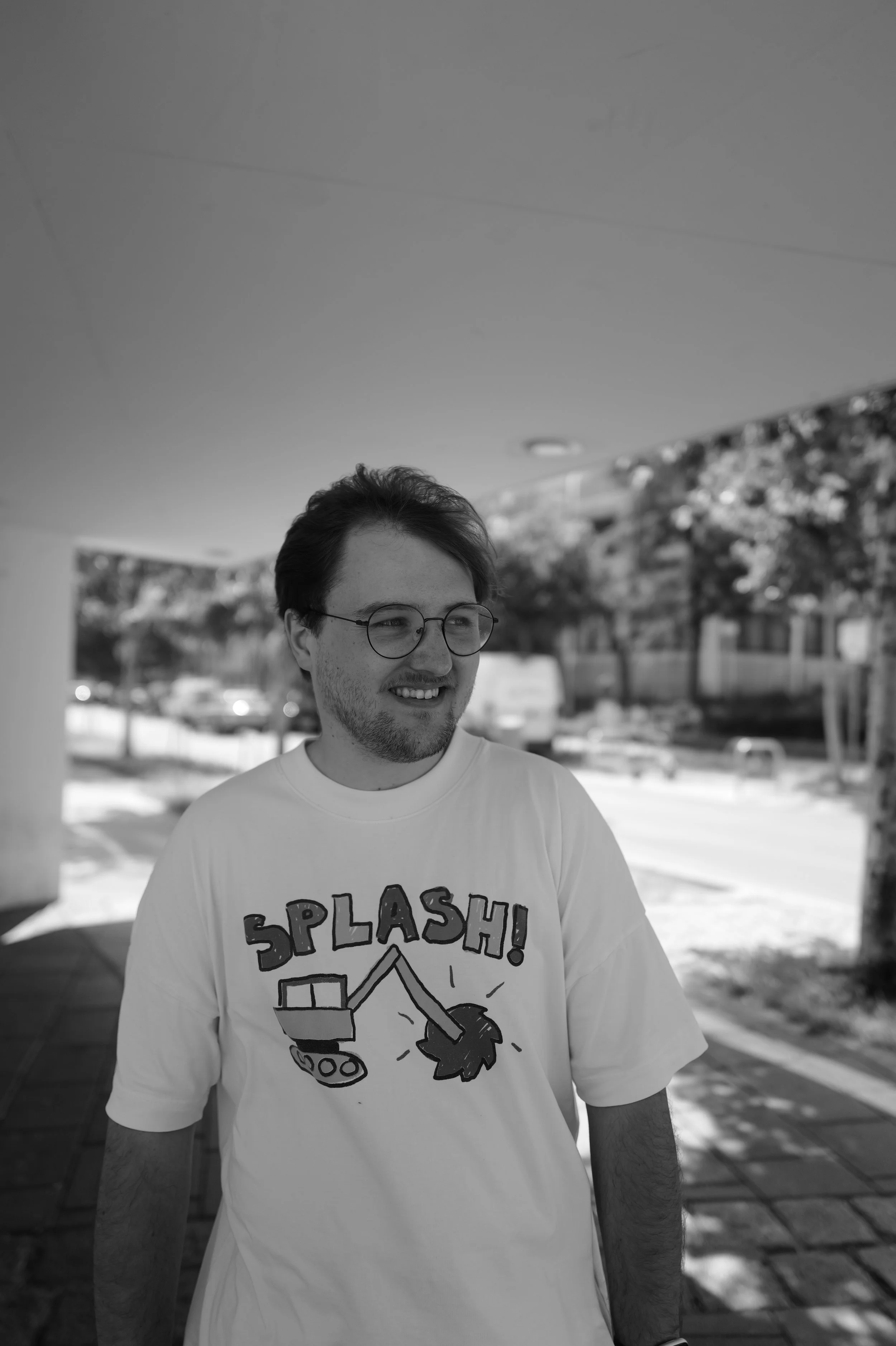 Junger Mann mit Brille und einem T-Shirt mit einem Spruch, der in einer städtischen Umgebung steht, im Freien, sonnig, schwarz-weiß Foto.