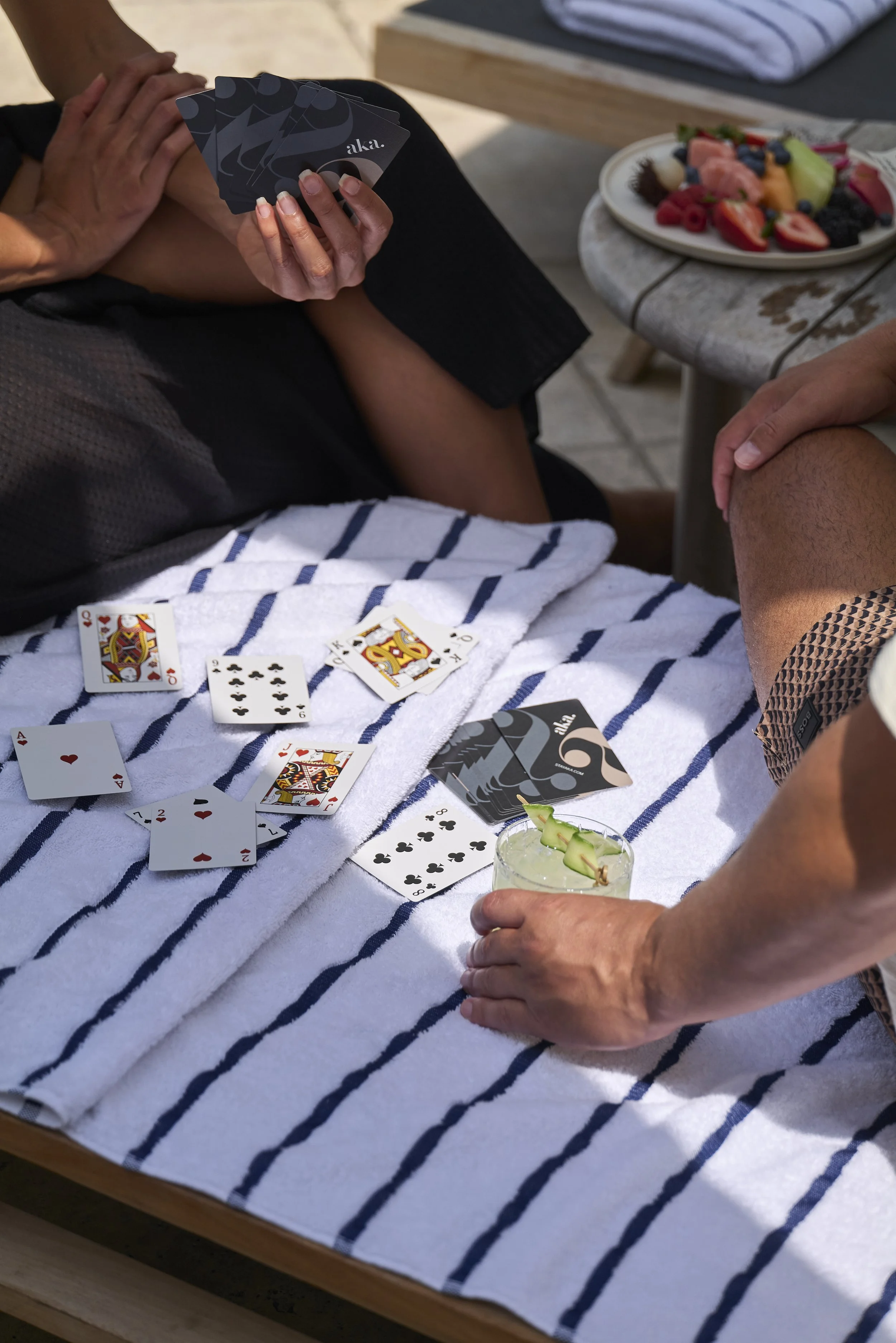 People playing a card game on a striped towel, with a plate of fruit and a drink on a small table nearby.