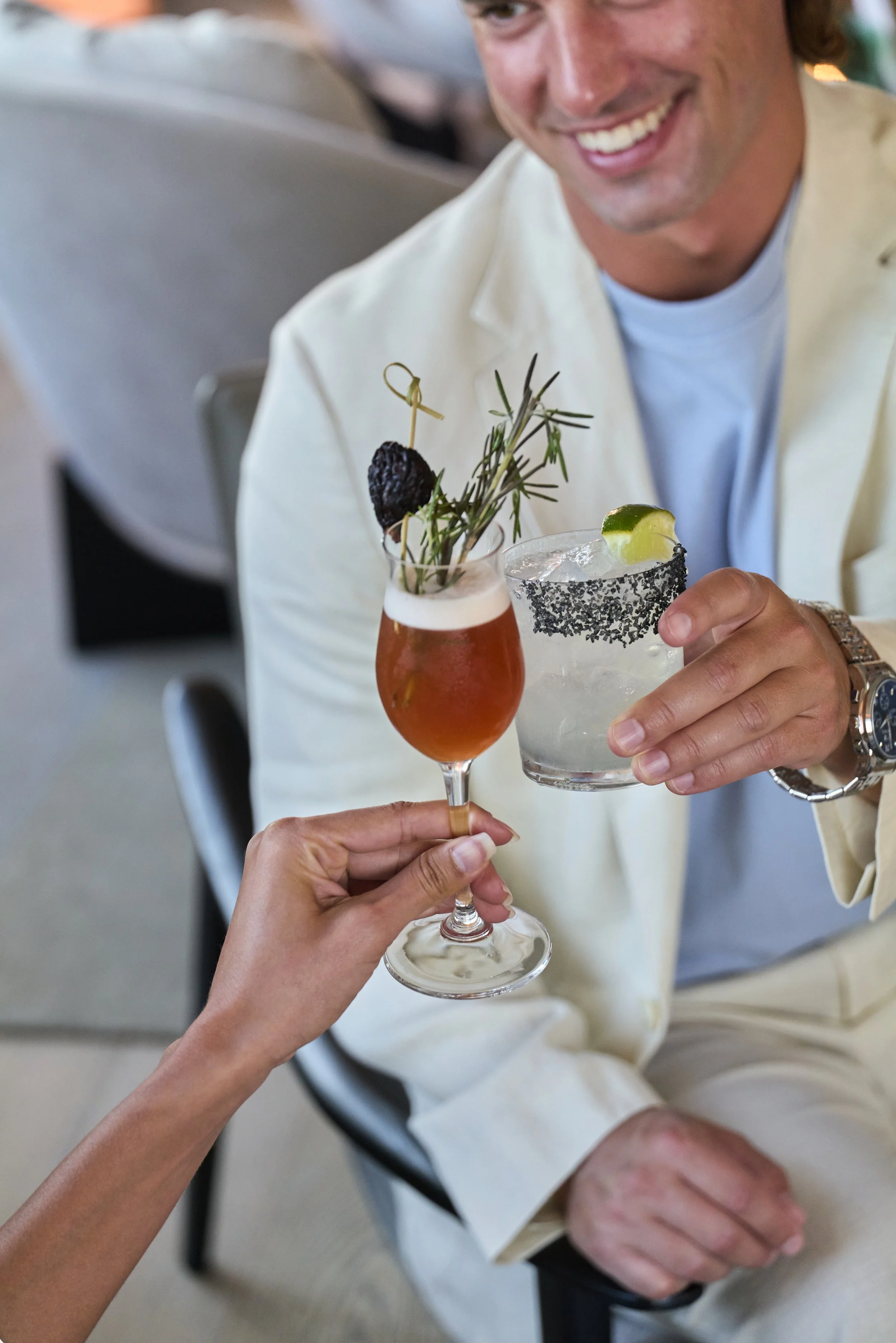 A man in a cream-colored suit smiling and holding a cocktail glass. Someone else is toasting him with a drink that has a lime wedge and garnishes, including rosemary and a blackberry. The setting appears to be a social gathering or celebration.
