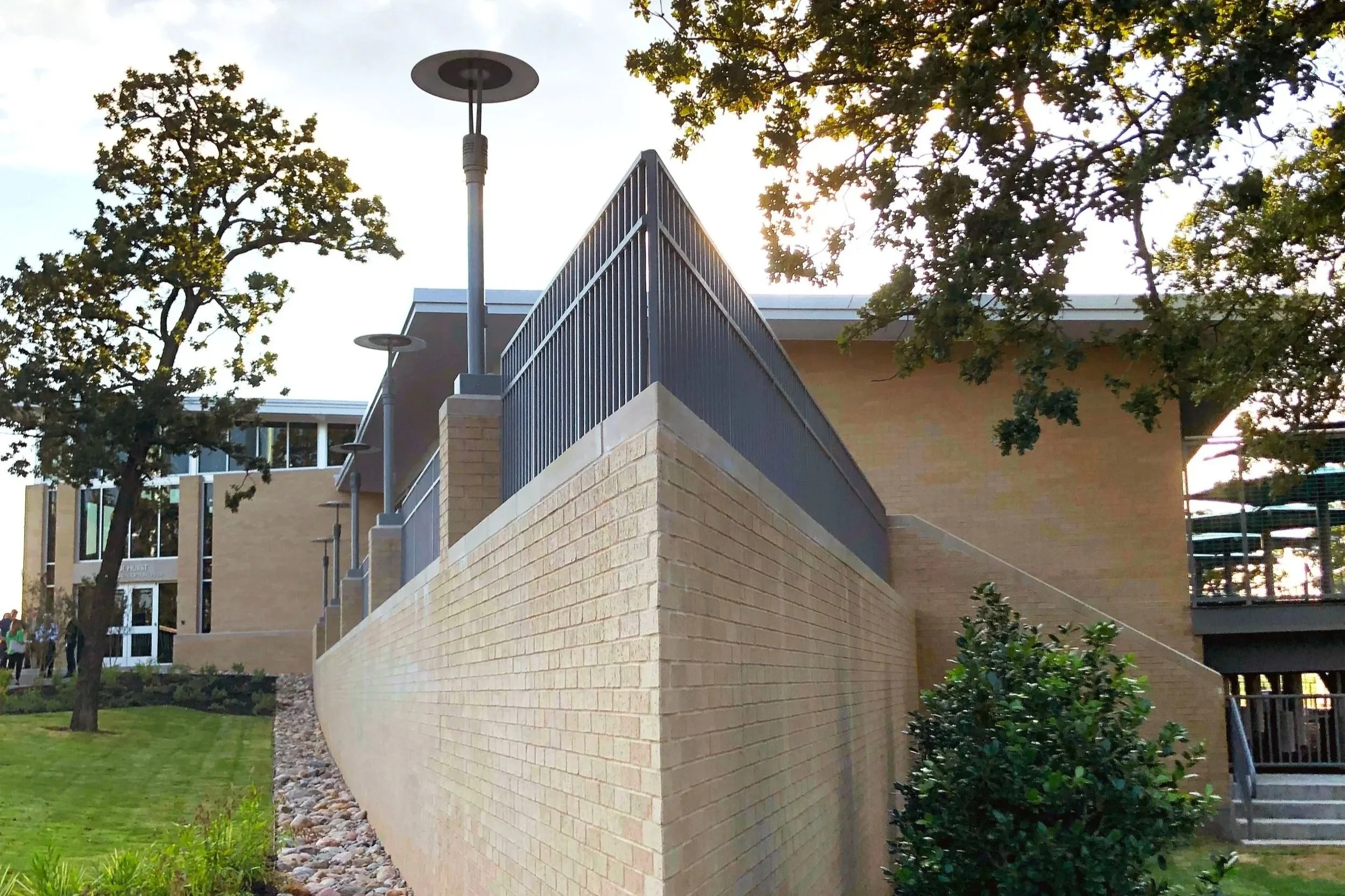 View of a modern building with a brick wall and metal railing, surrounded by green trees and grassy area, with sunlight peeking through the leaves.