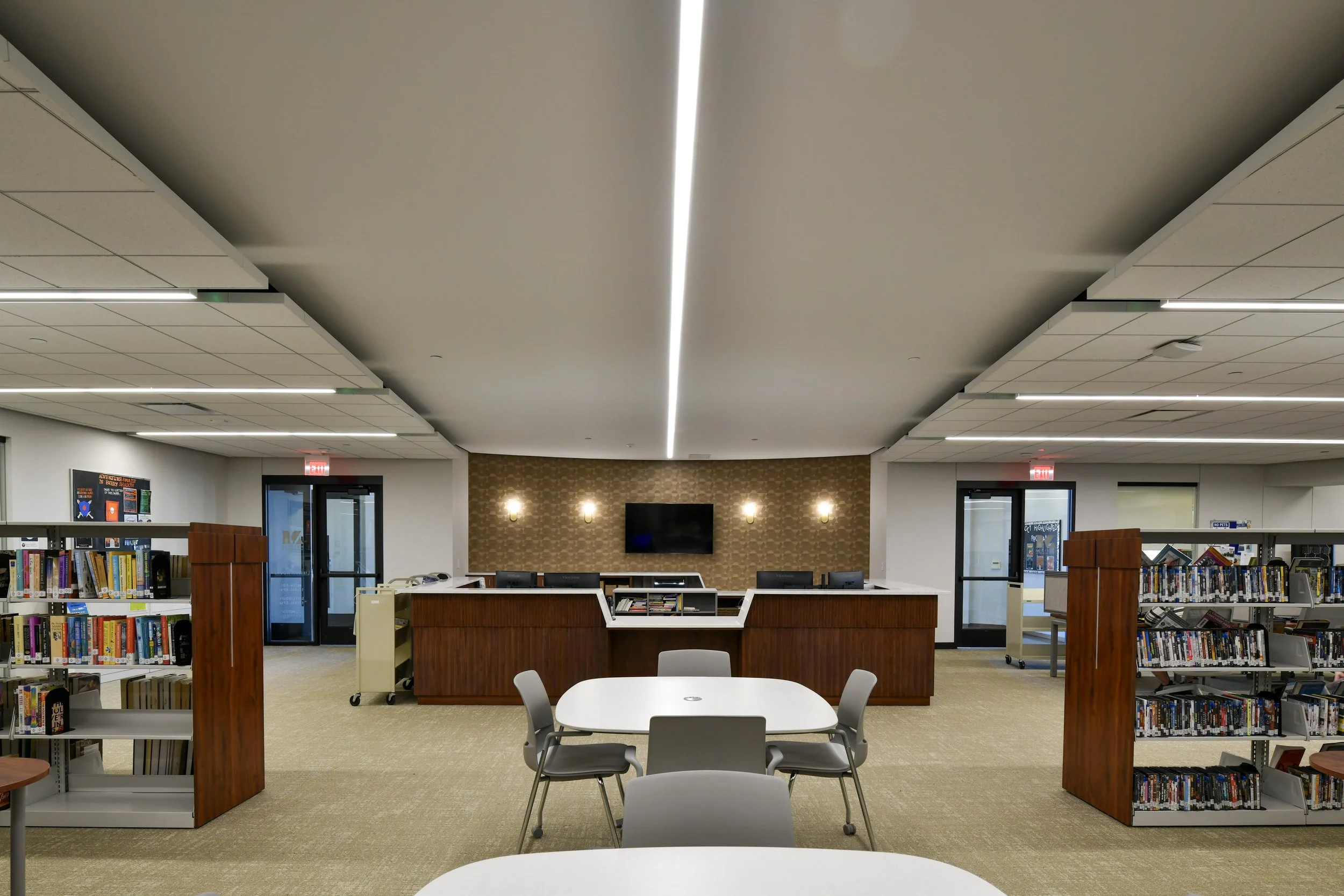 Interior view of a modern library or community center with bookshelves, tables, chairs, a TV, and decorative lighting on the wall.