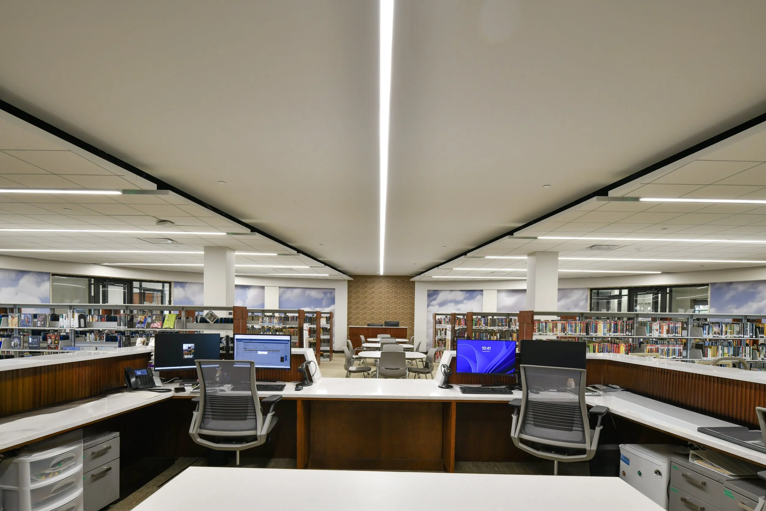 Interior view of a modern library with white desks, computer monitors, and shelves filled with books, illuminated by linear ceiling lights.