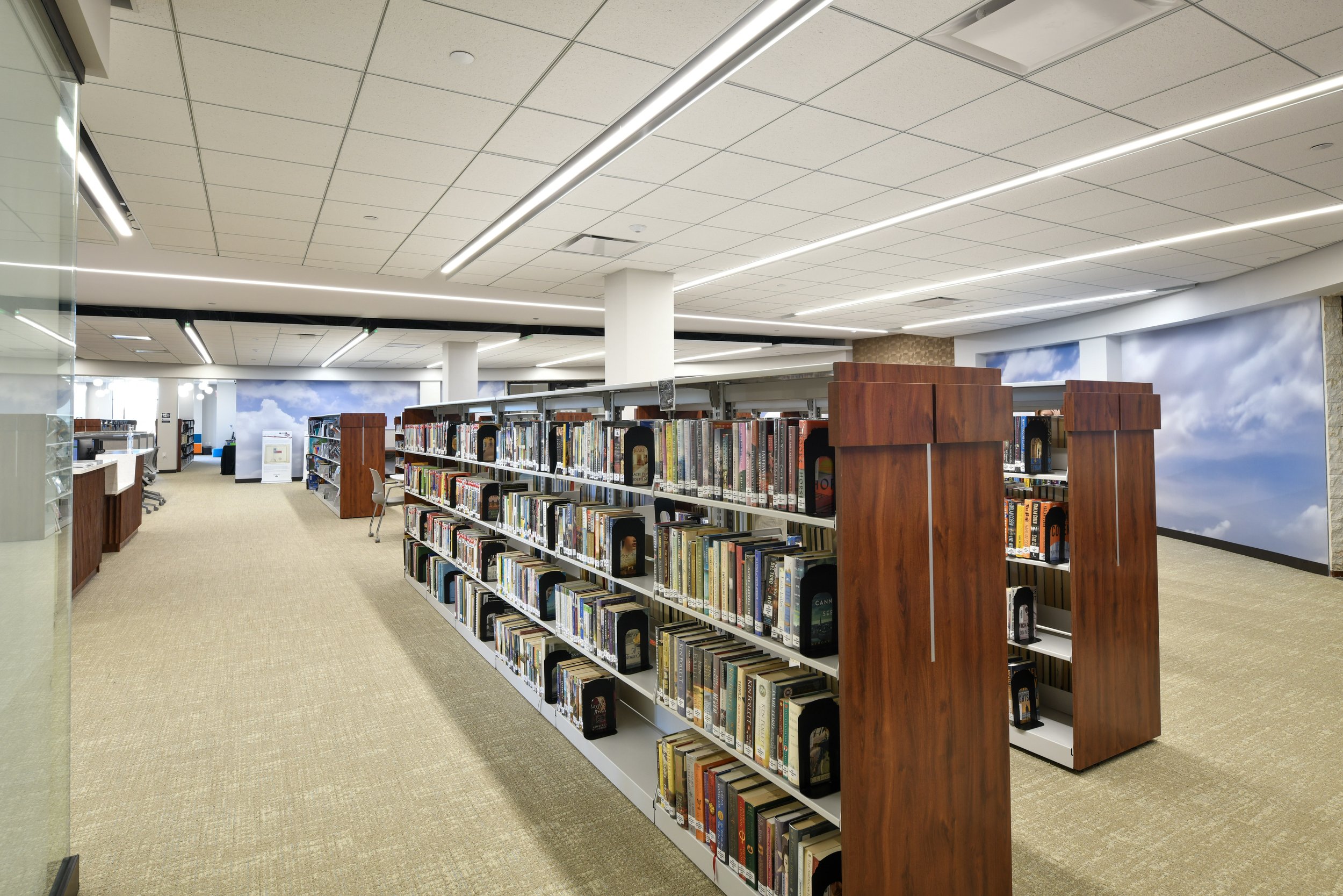 A view of a modern library with shelves full of books, a beige carpeted floor, white ceiling with long linear lighting, and a large mural of a cloudy sky on the wall.