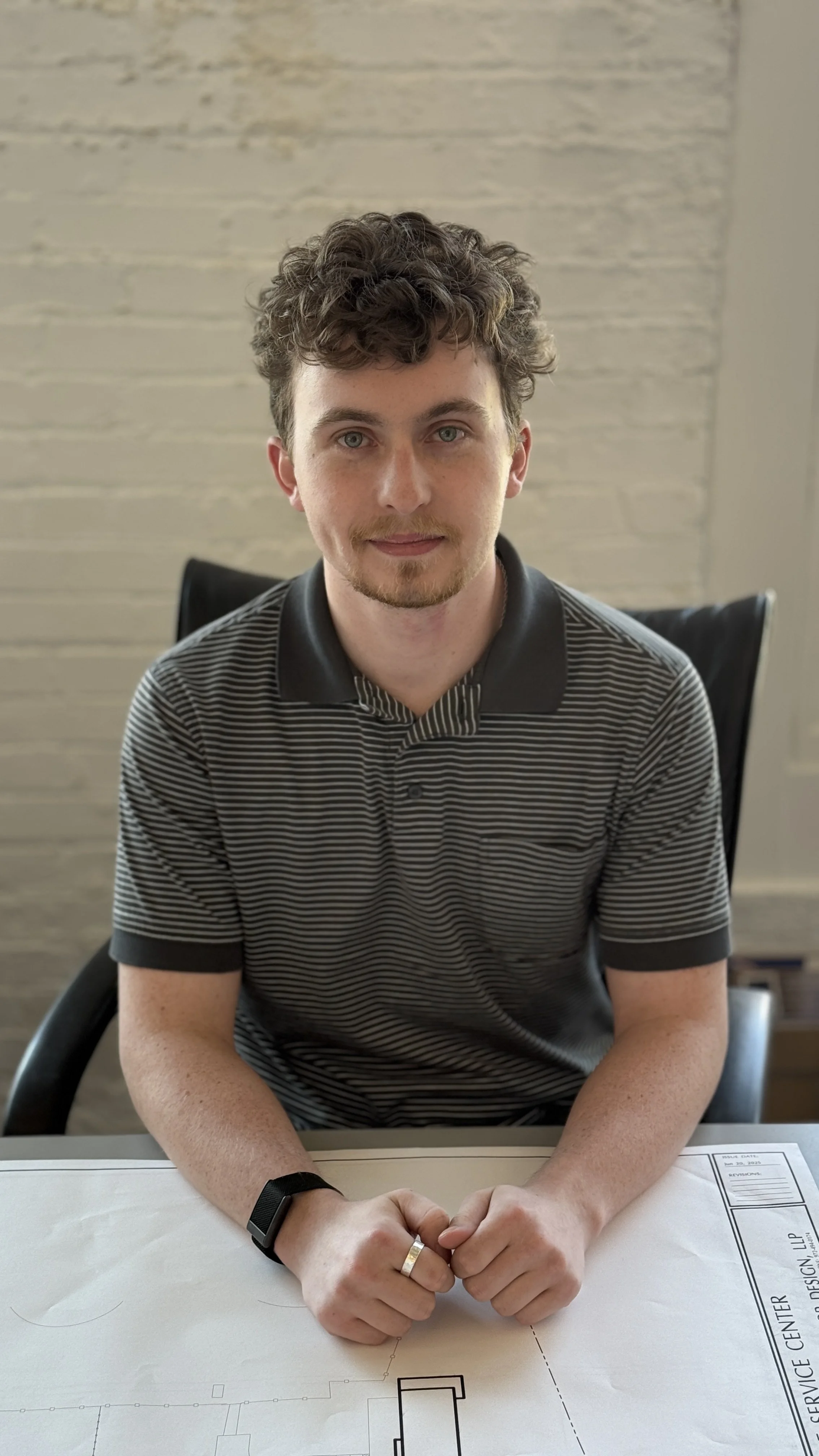 A young man with curly hair and a goatee, wearing a striped polo shirt, sitting at a table with hands clasped, in a room with a textured wall background.