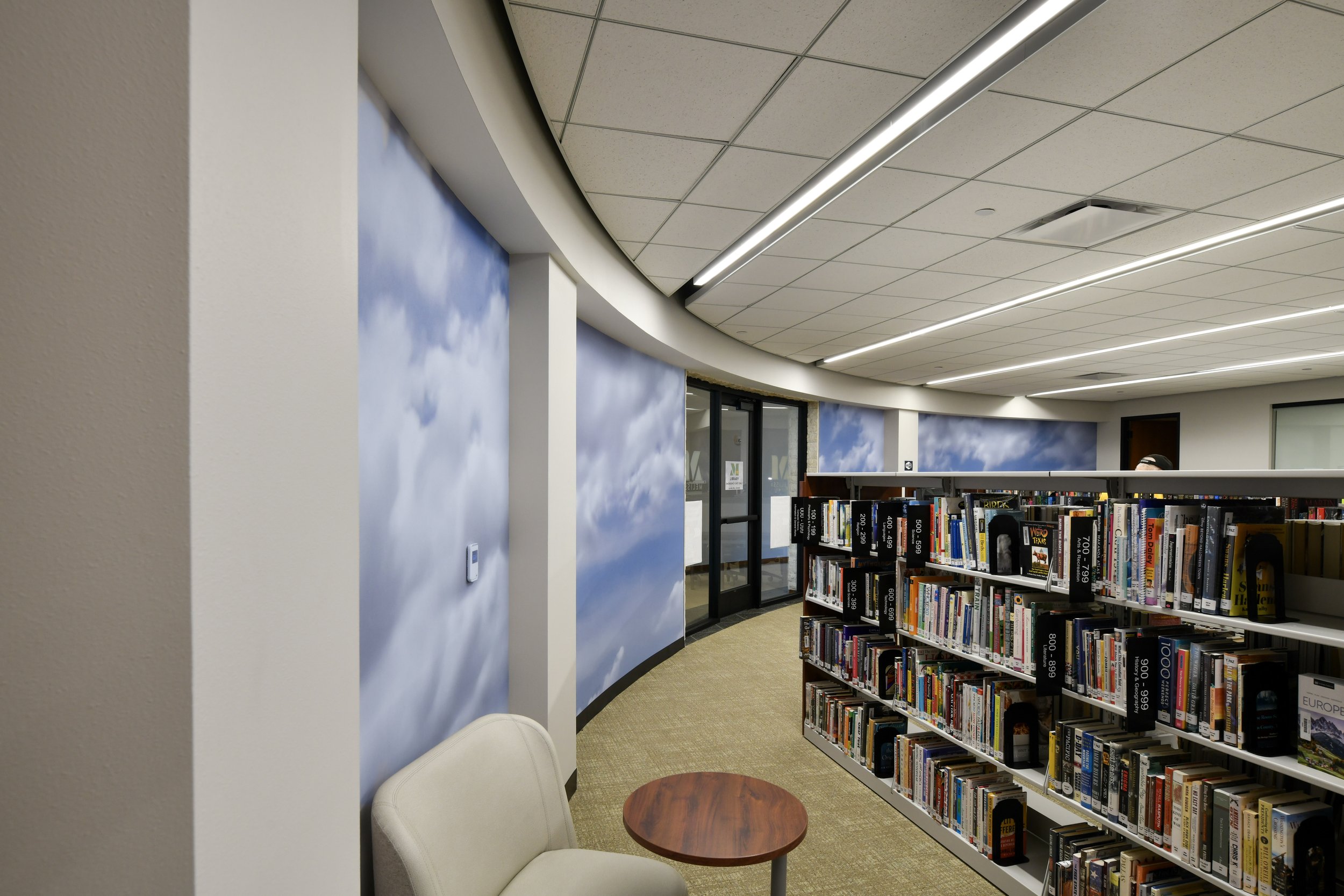 Interior of a library with bookshelves, a small round table, chairs, and a mural of clouds on the wall.