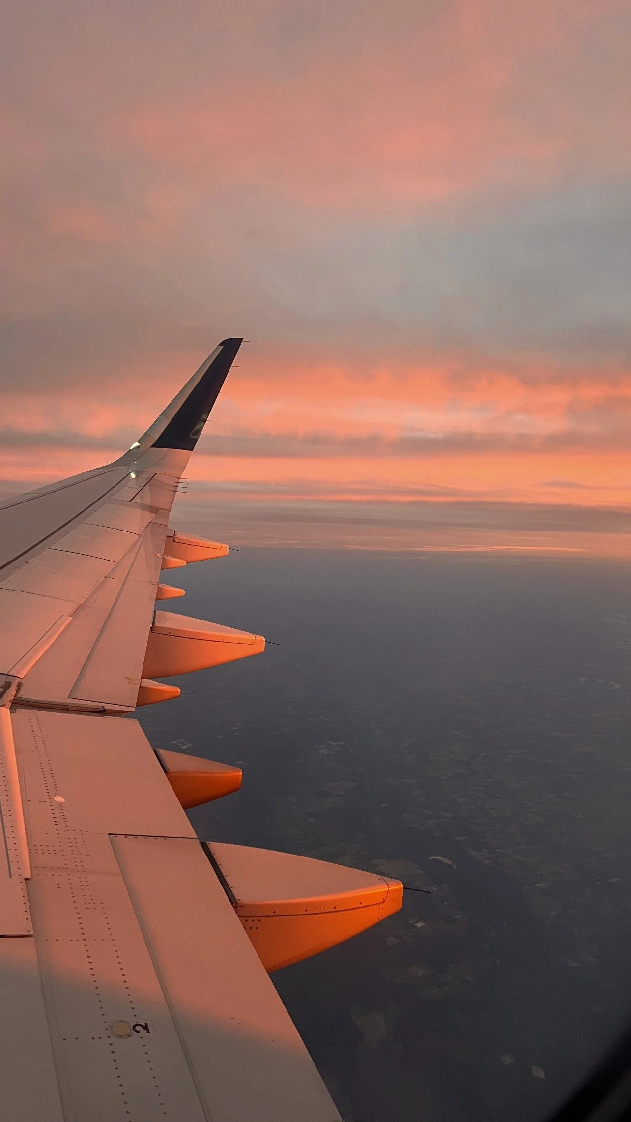 View from airplane window showing airplane wing against sunset sky with pink, orange, and purple clouds.