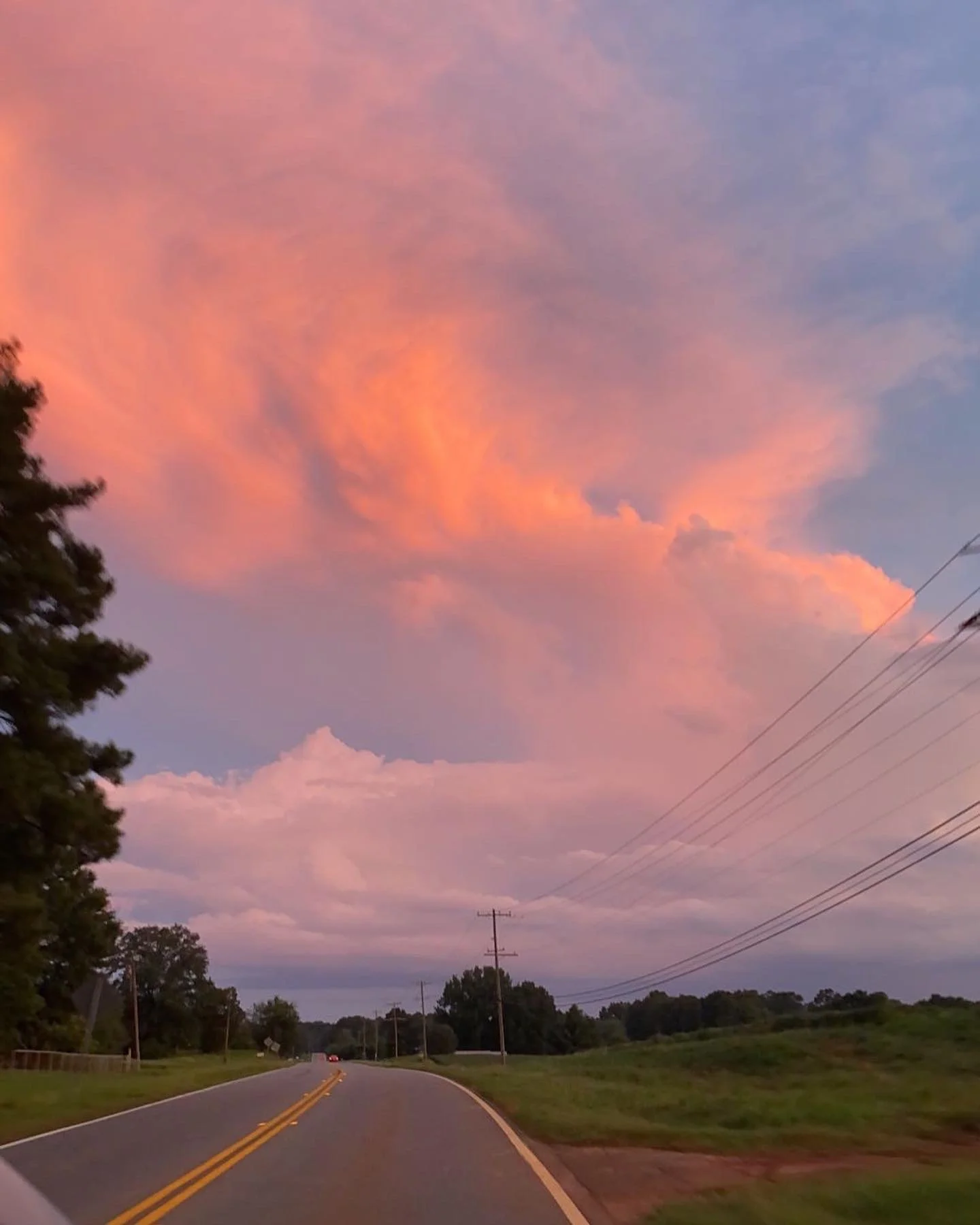 A rural road at sunset with a pink and purple sky, trees on the left, power lines on the right, and a car in the distance.