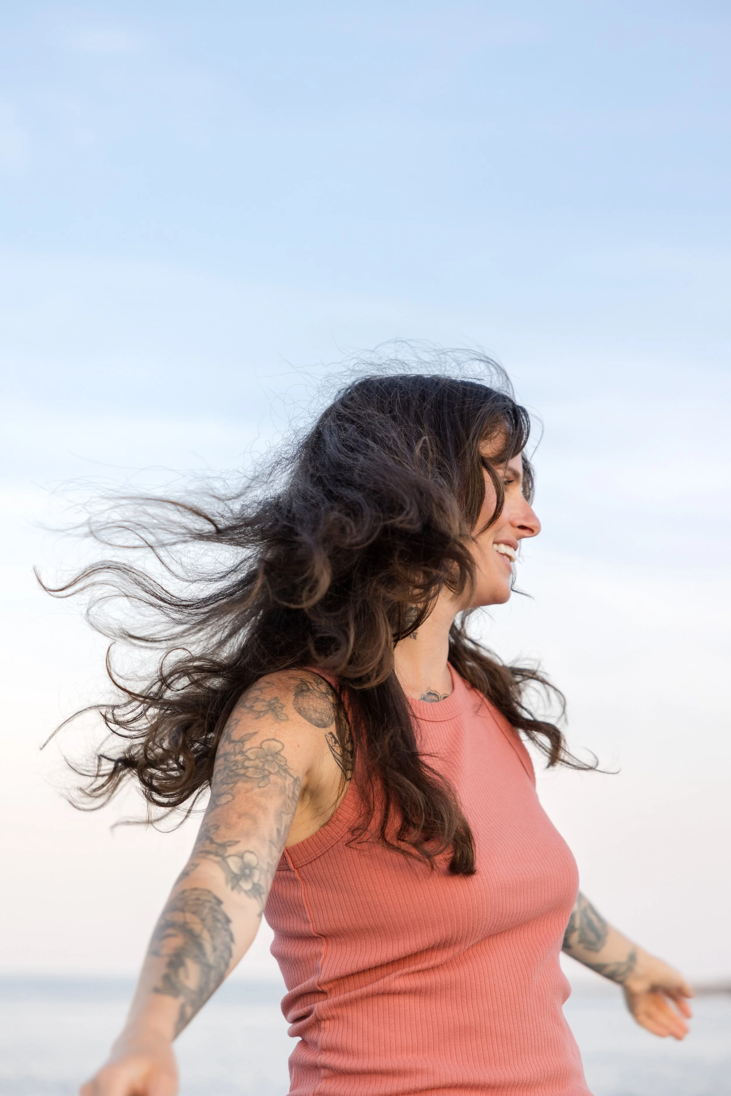 A woman with long dark hair and tattoos on her arms is smiling and looking to the side while standing outdoors. The background shows a cloudy sky and a hint of the beach.