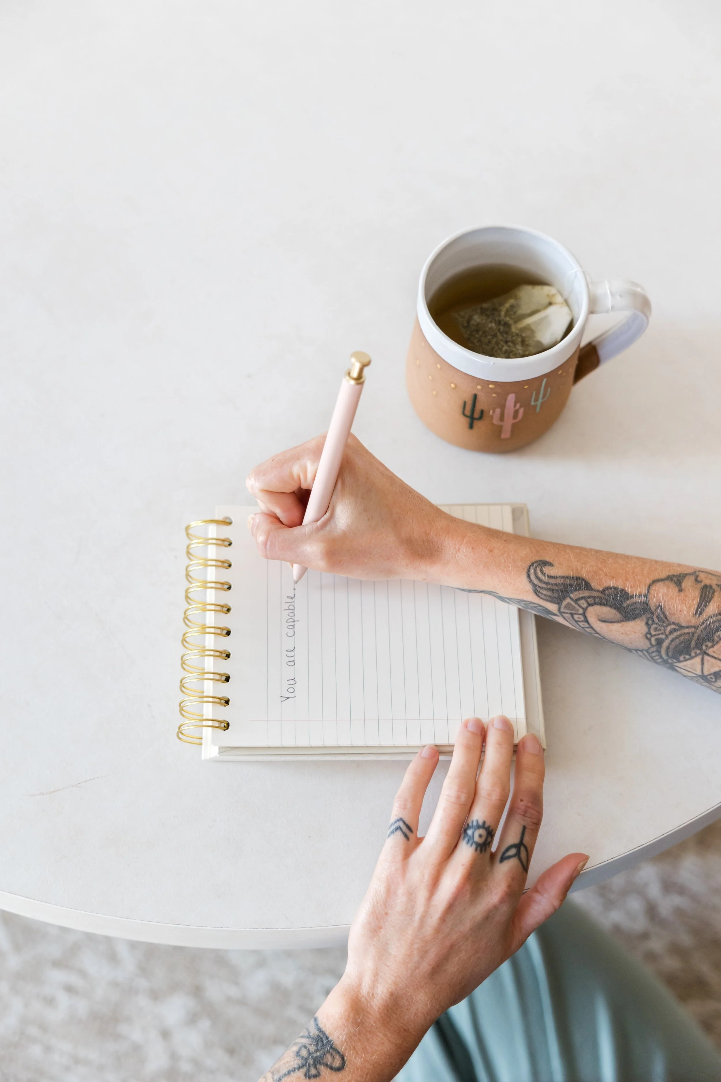 A person with tattoos writing in a spiral notebook at a white table with a tea mug nearby.