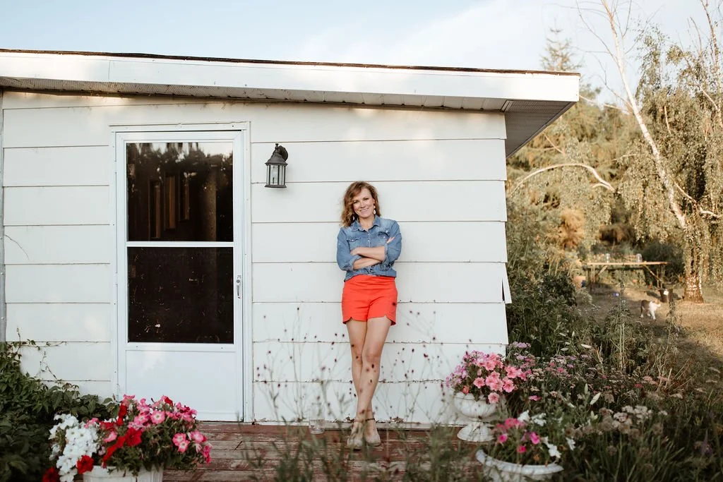 Woman standing and smiling against a white house wall, wearing a denim jacket and red shorts, with potted flowers in front and trees in the background.