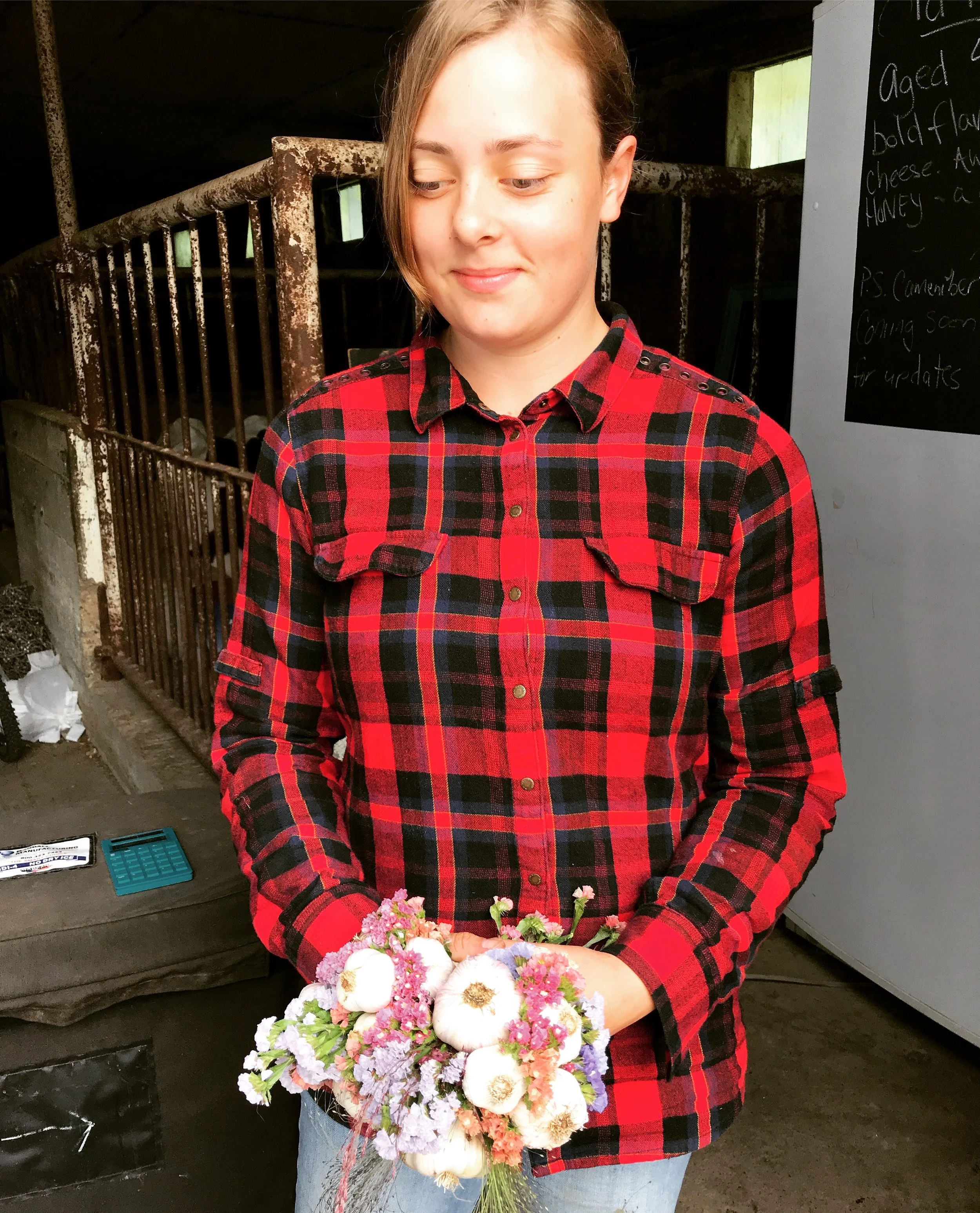 A young woman in a red and black plaid shirt holding a bouquet of pink, white, and purple flowers, standing indoors near a chalkboard and rusted metal bars.