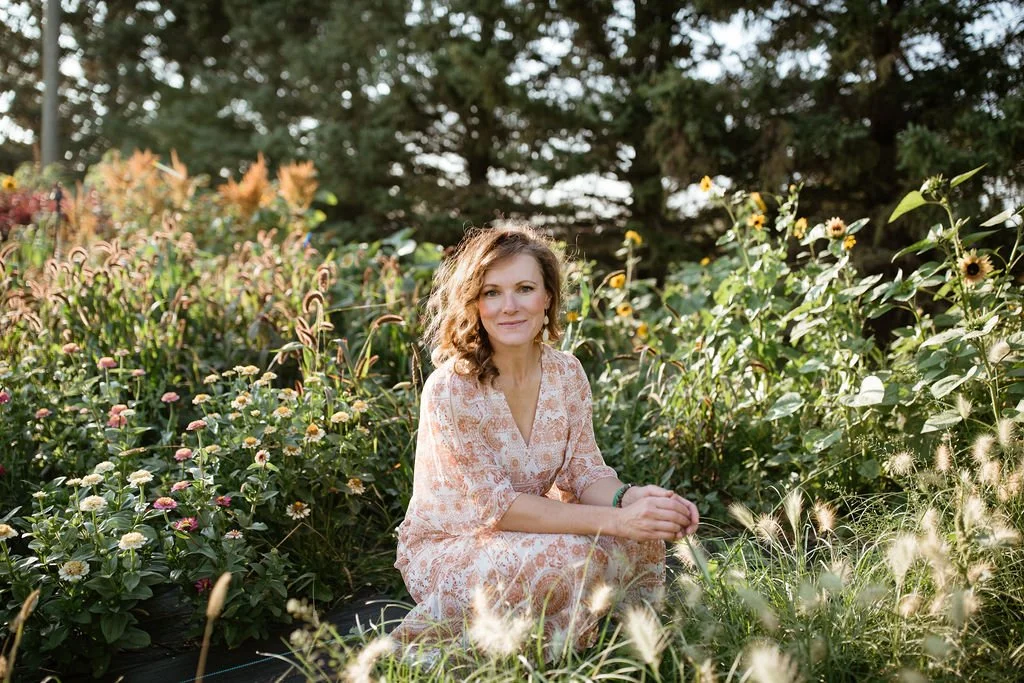 A woman with wavy brown hair sitting among various flowering plants and greenery in a garden.