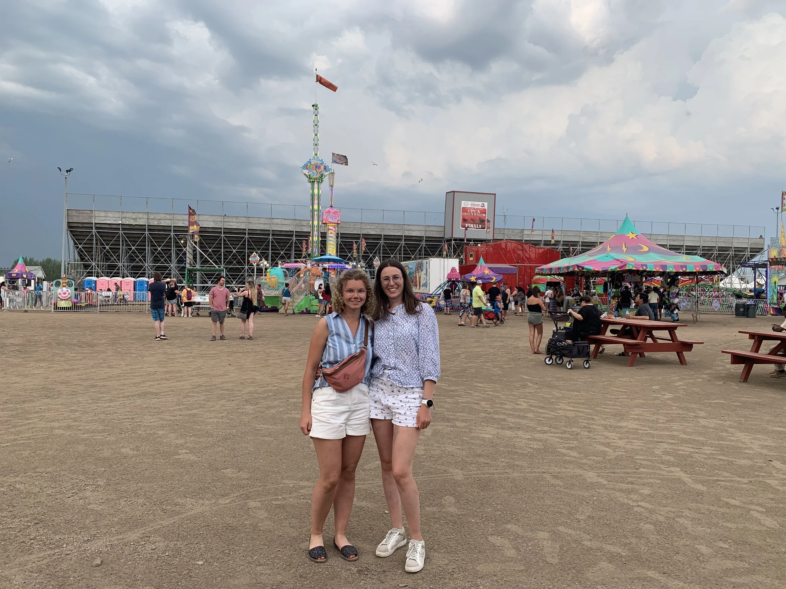 Two young women smiling at a fairground with rides, colorful tents, and people in the background on a cloudy day.