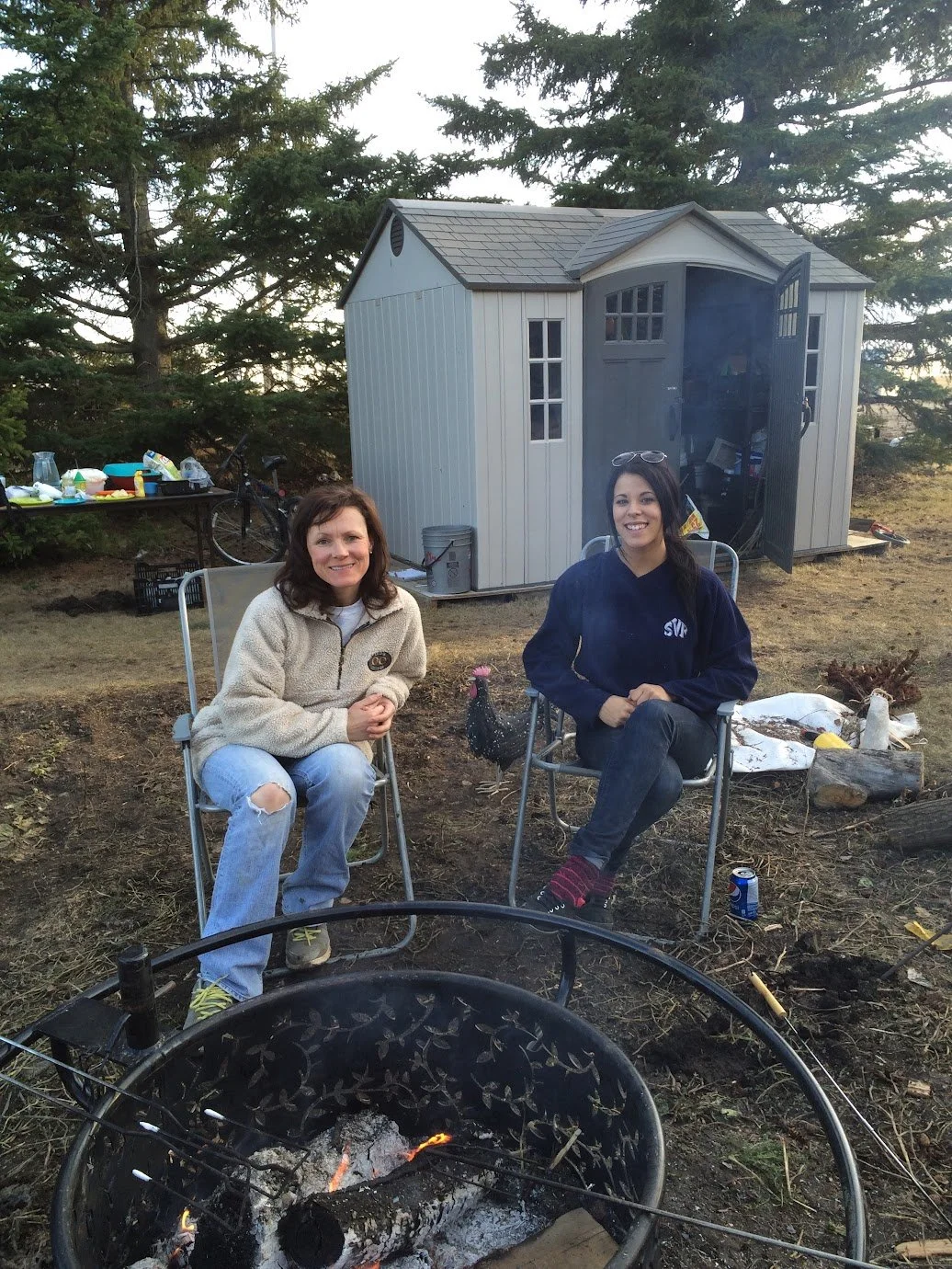 Two women sitting on outdoor chairs around a campfire with a shed, trees, and camping supplies in the background.