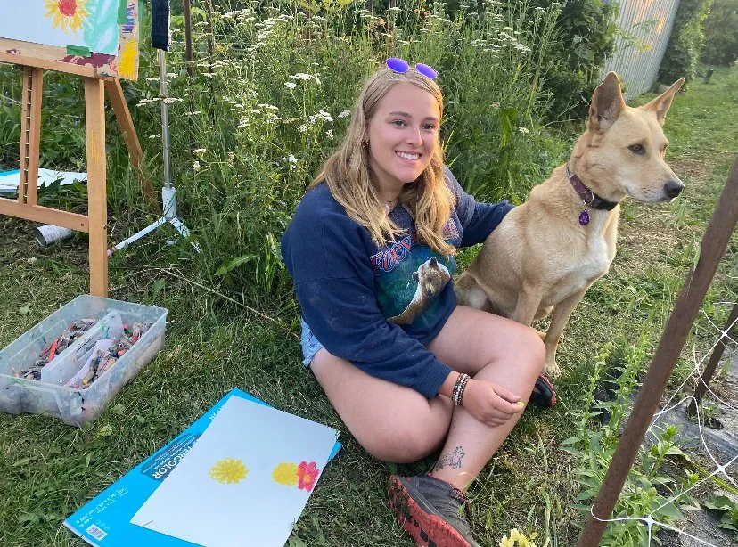 A young woman with blonde hair, wearing sunglasses, a blue jacket with a space-themed design, and shorts, is sitting on the grass outdoors next to a tan and white dog with pointy ears, on a sunny day. She is smiling and looking at the camera. There are painting supplies and paper nearby, with a colorful painting on an easel in the background.