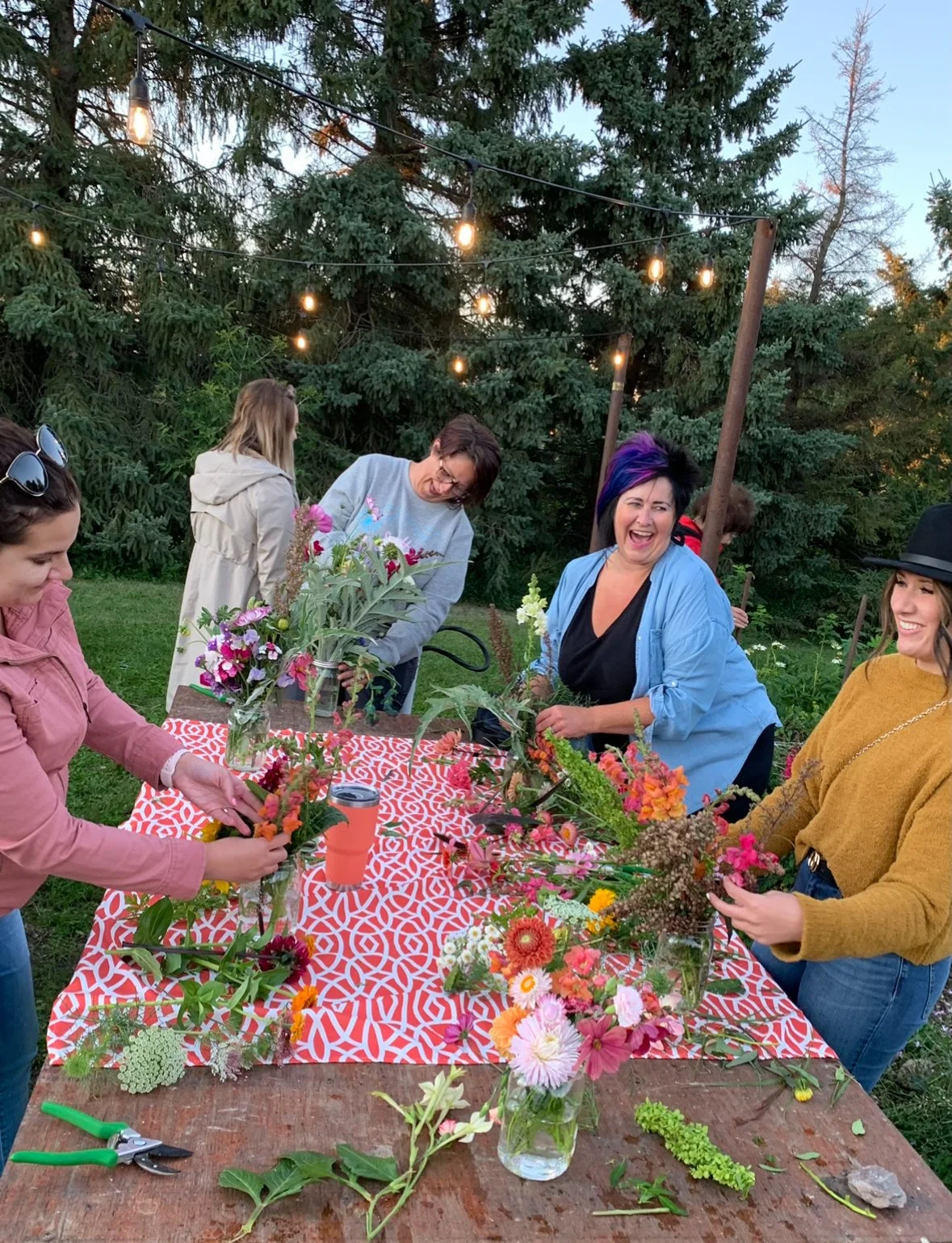 Group of women arranging colorful flowers in mason jars on a decorated outdoor table at sunset.
