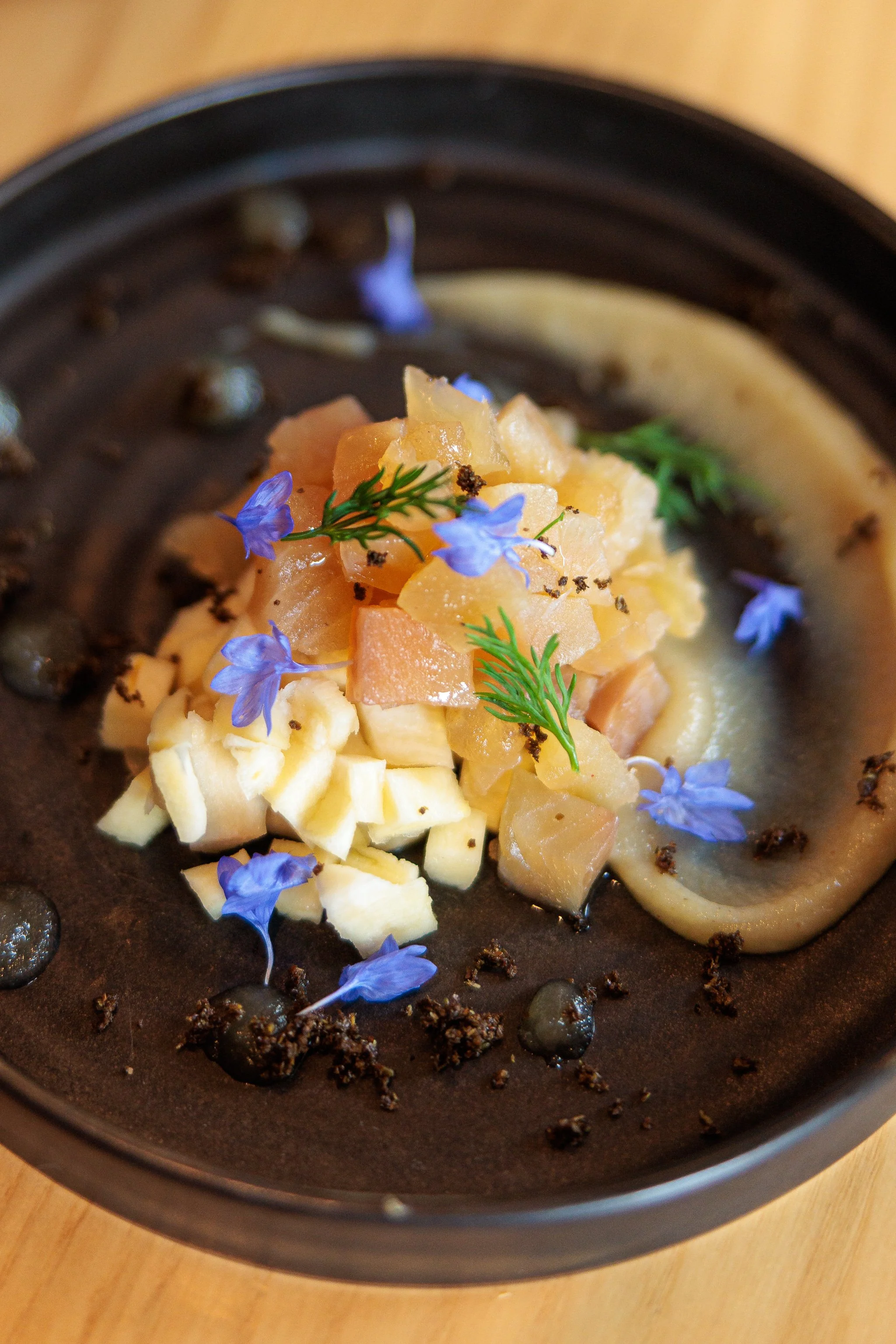 A gourmet dish featuring diced vegetables, edible flowers, and herbs on a black plate.