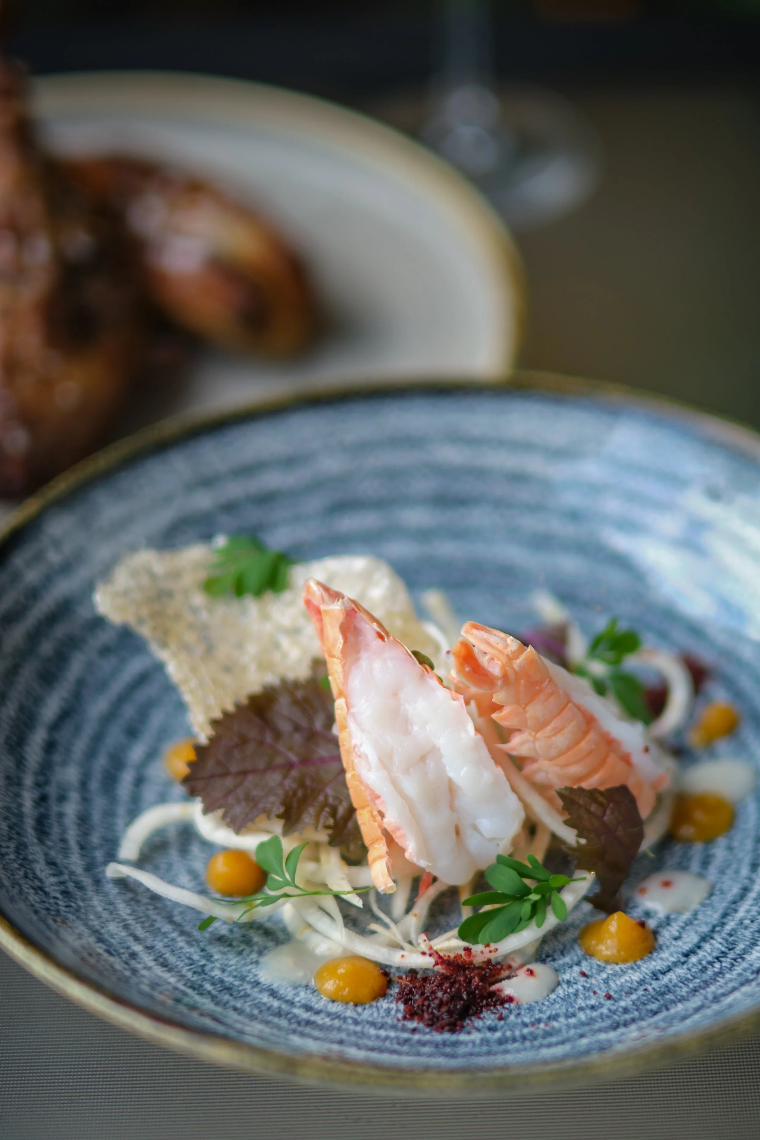 A gourmet dish featuring lobster tail slices, garnished with herbs, accompanied by colorful sauces and edible leaves on a blue ceramic plate.