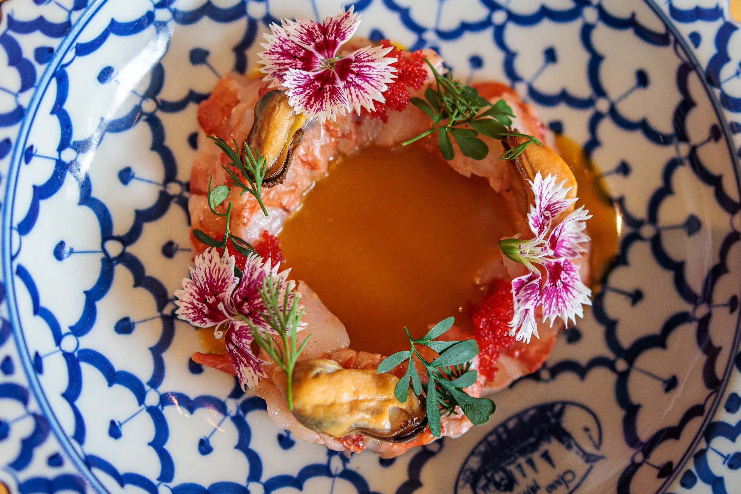 A gourmet seafood dish garnished with edible flowers and microgreens, presented on a blue and white patterned plate.
