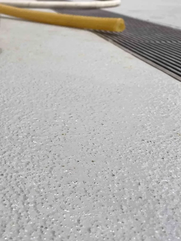 Close-up of a textured white surface, likely flooring, with a black grated drain and a yellow object near the top of the image.