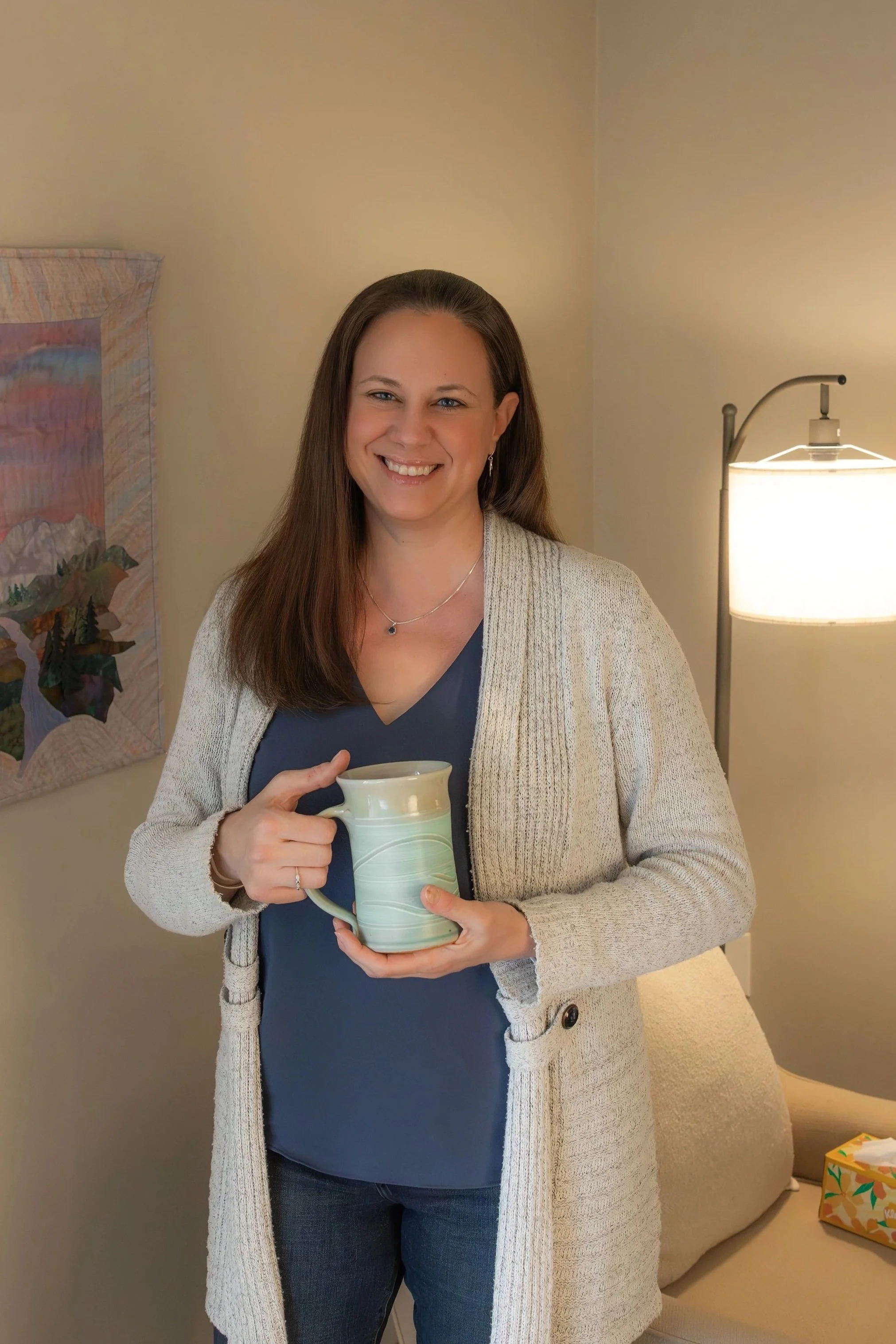 A woman with long brown hair, smiling, holding a ceramic mug, standing in a cozy office with a lamp, artwork, and a tissue box on the side.