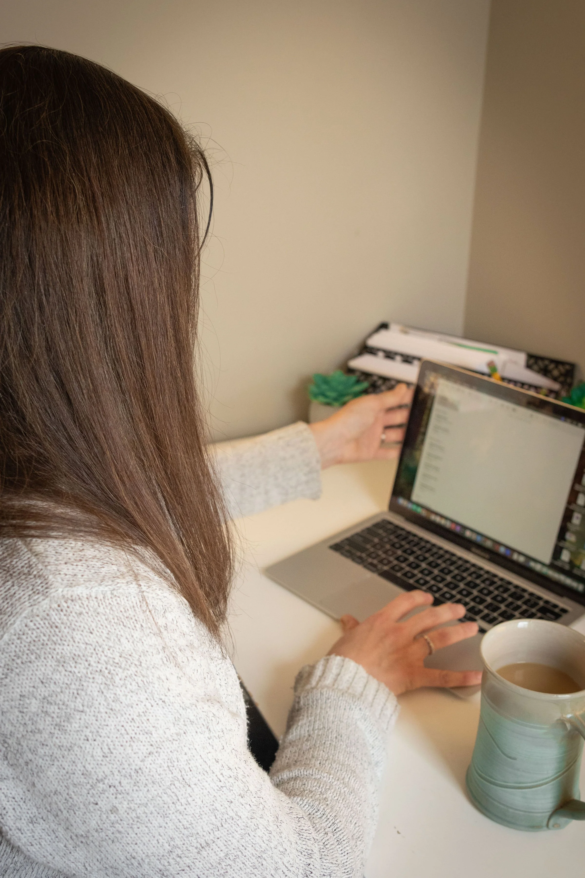 A woman with brown hair sitting at a desk, working on a laptop, with a cup of coffee and a ceramic vase nearby.
