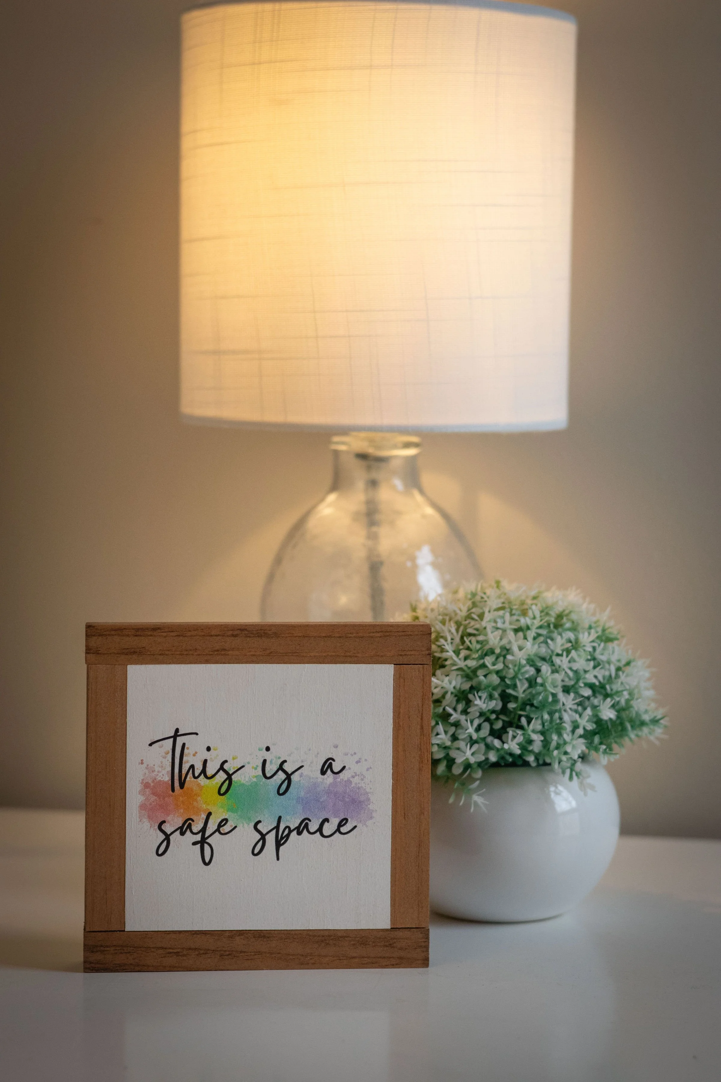 A framed sign with rainbow watercolor background reading 'This is a safe space,' placed on a white surface next to a white vase with small green and white flowers, with a glowing table lamp in the background.