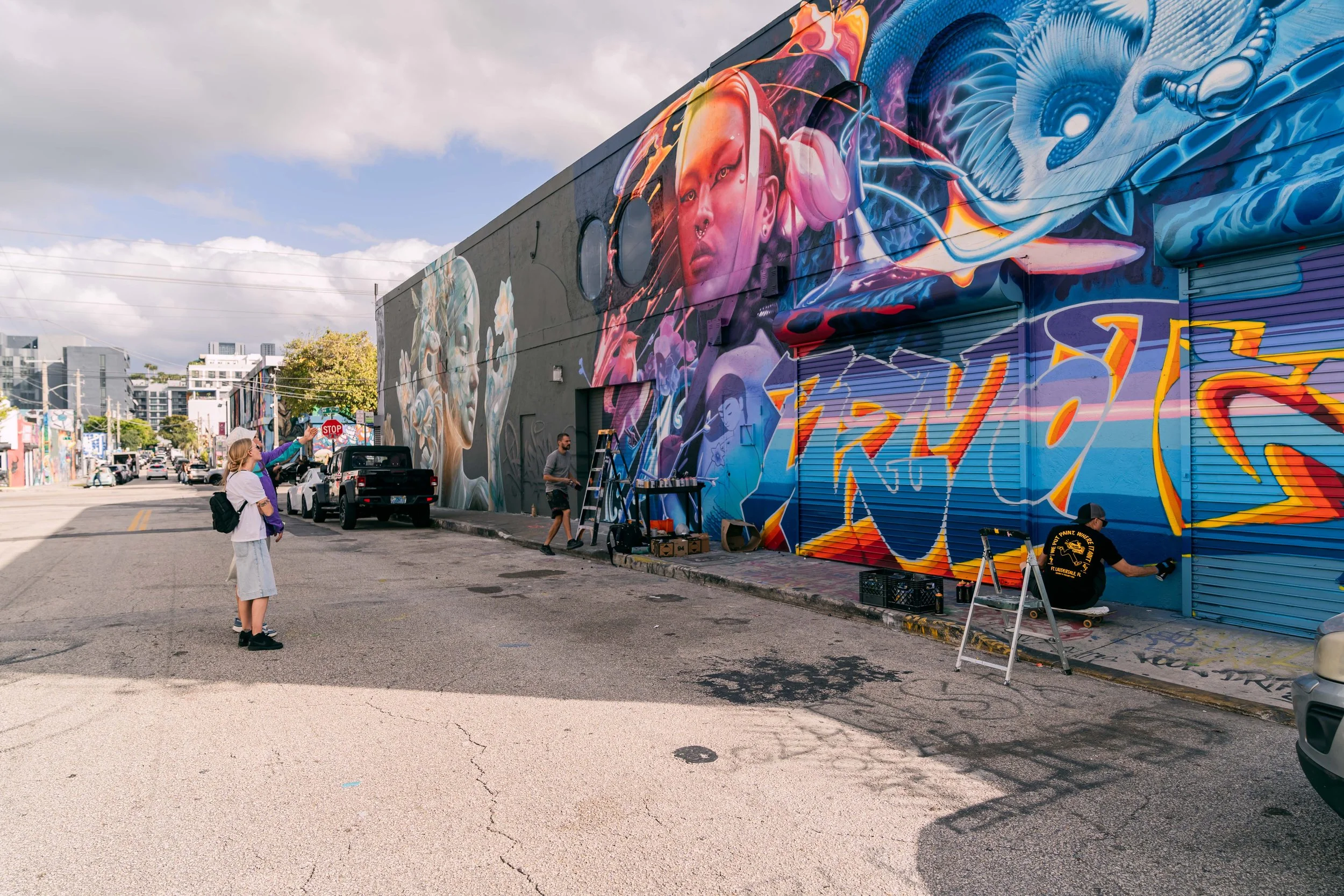 Artists creating colorful mural art on the exterior wall of a building in an urban area, with a few people watching and working.