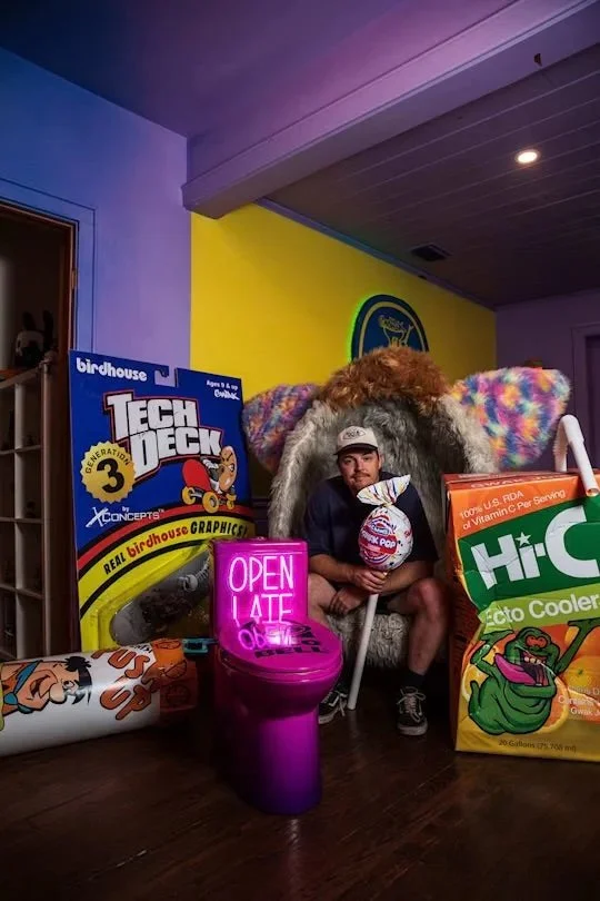 A man sitting in a large, colorful chair that resembles a crab or lobster, surrounded by giant objects such as a toy birdhouse, a neon 'Open Late' sign, a giant bag of Hi-C Ecto Cooler, and a figure of Popeye. The background features a yellow and purple wall with a logo or decoration.