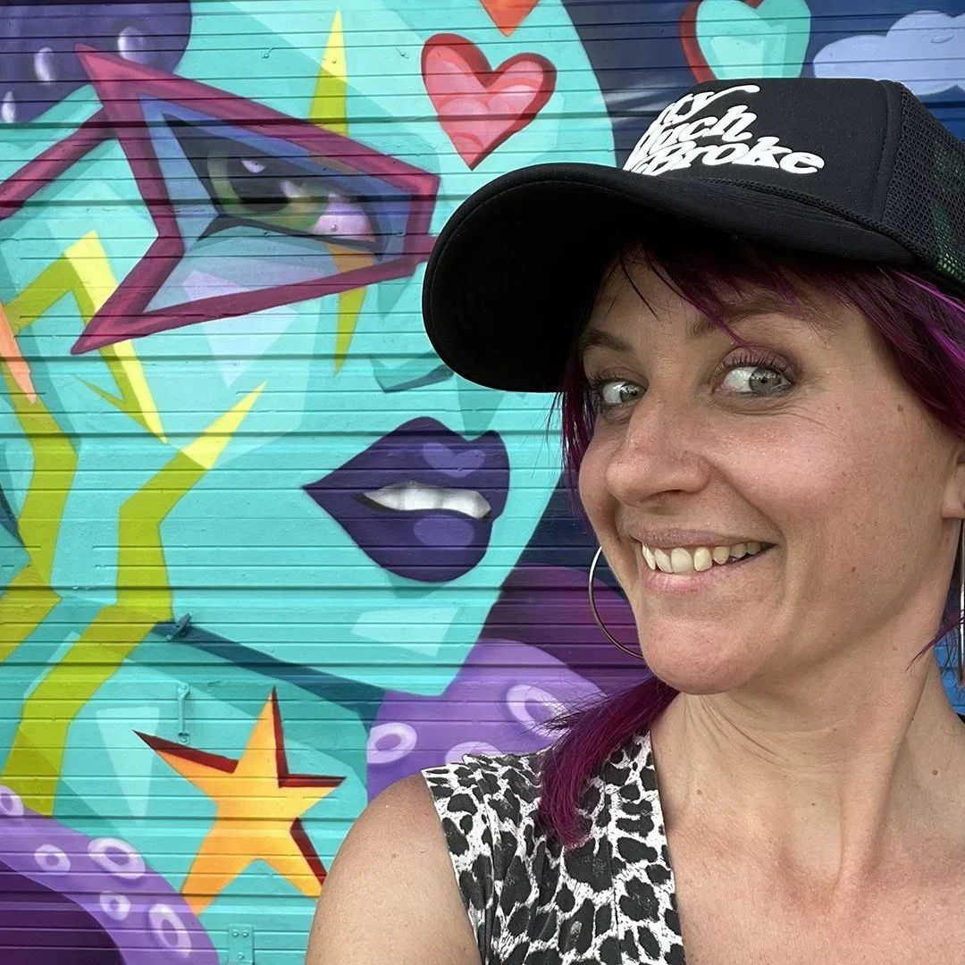 Artist Skela with purple hair and a black cap standing in front of a colorful graffiti mural featuring a stylized face of a woman, hearts, stars, and abstract shapes.