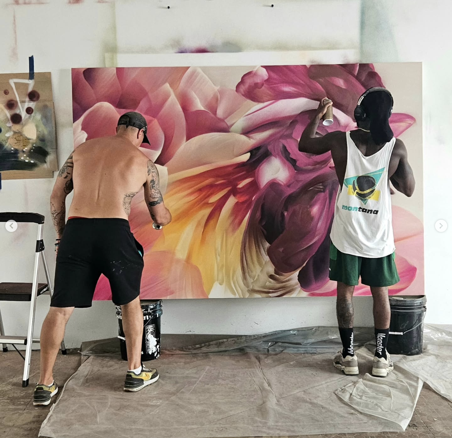 Two men creating a large floral graffiti art on the wall. One man is shirtless with tattoos, wearing black shorts and sneakers. The other is wearing a white tank top, green shorts, and a black balaclava, spray painting the mural. The mural features abstract, swirling pink, purple, and yellowflower-like shapes.