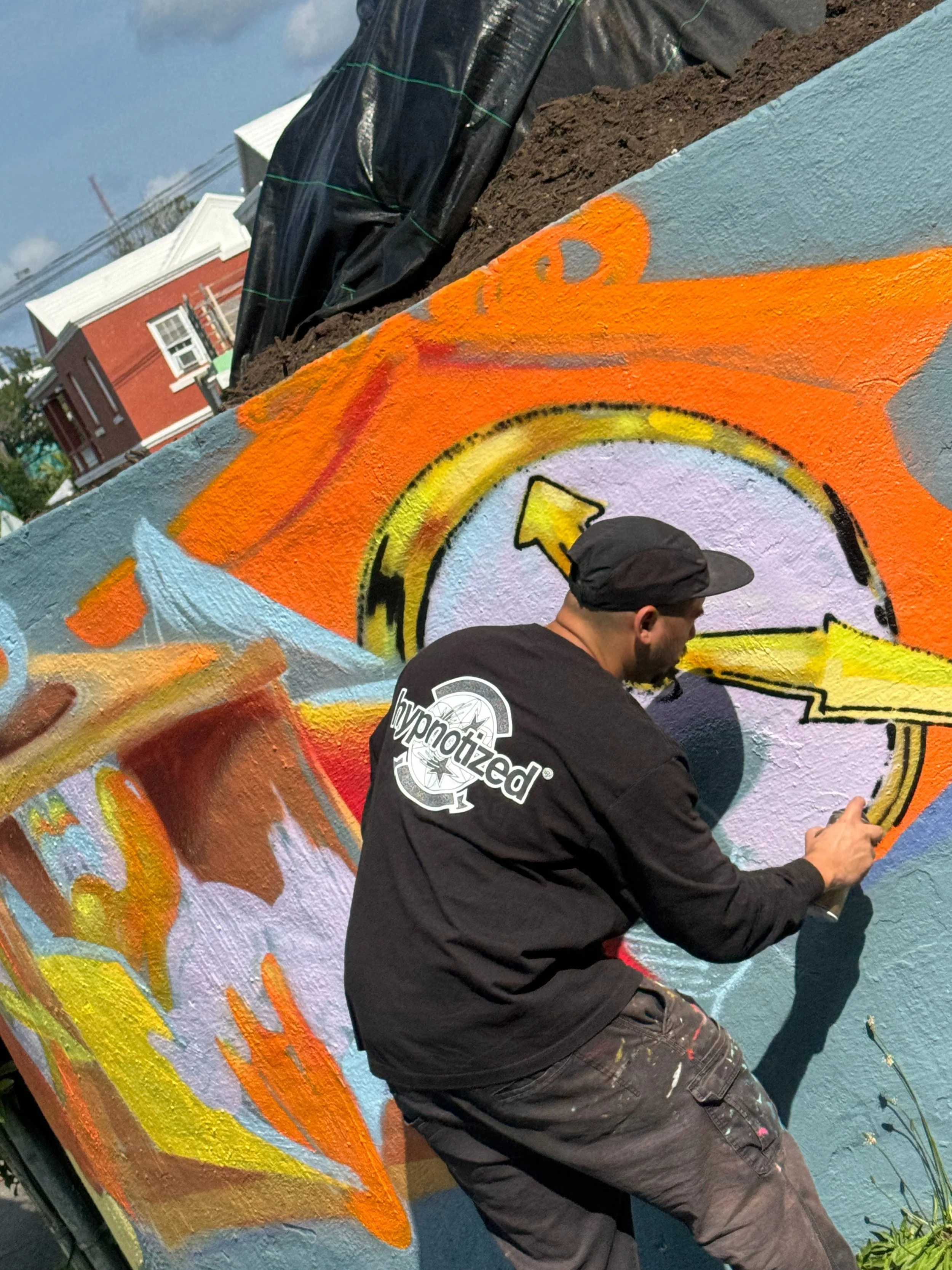 A man painting colorful graffiti on a wall outdoors, with a nearby building visible in the background.