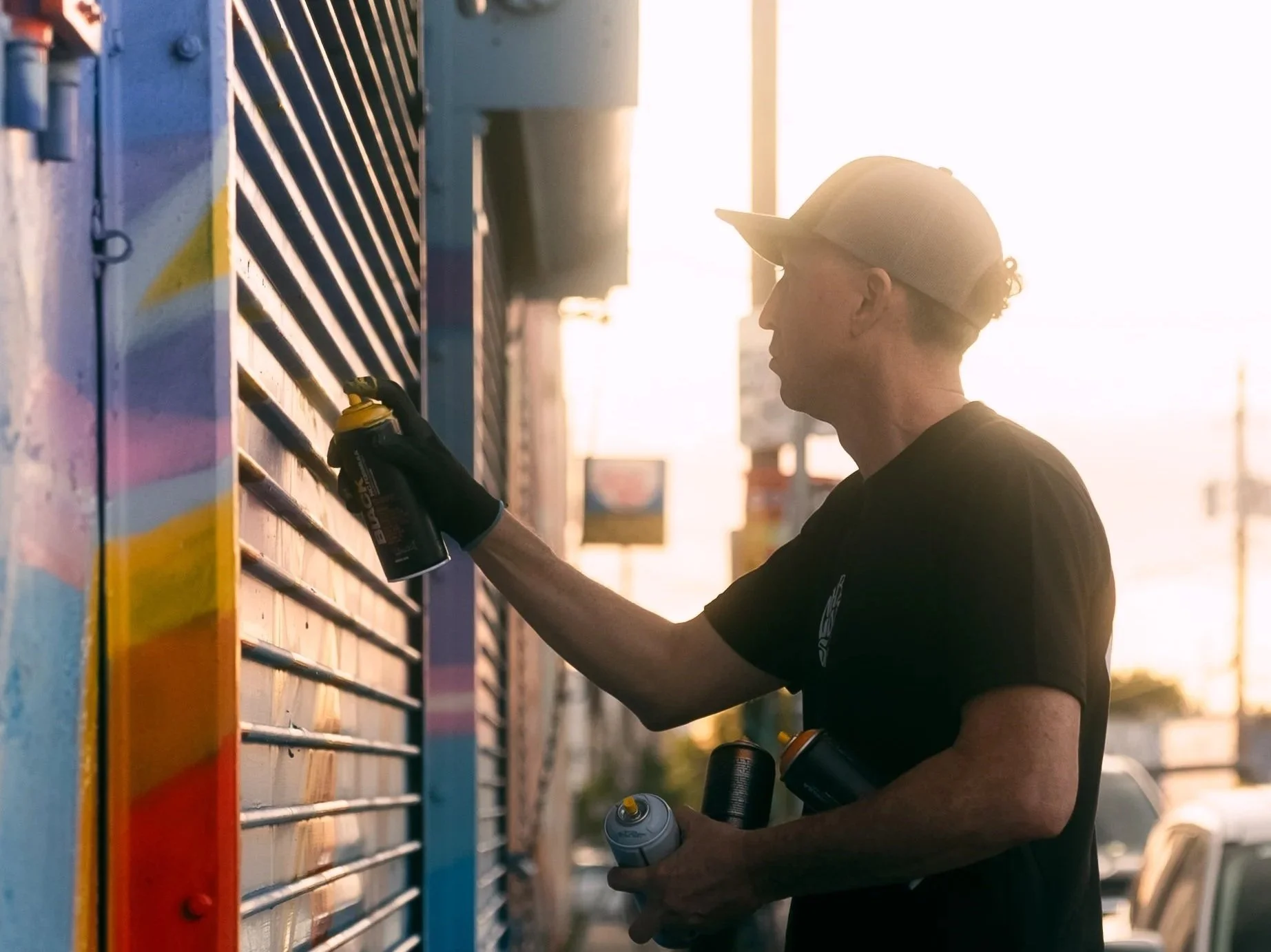 A man in a black t-shirt, shorts, and a cap spray paints colorful graffiti on a metal shutter in an urban setting during sunset.
