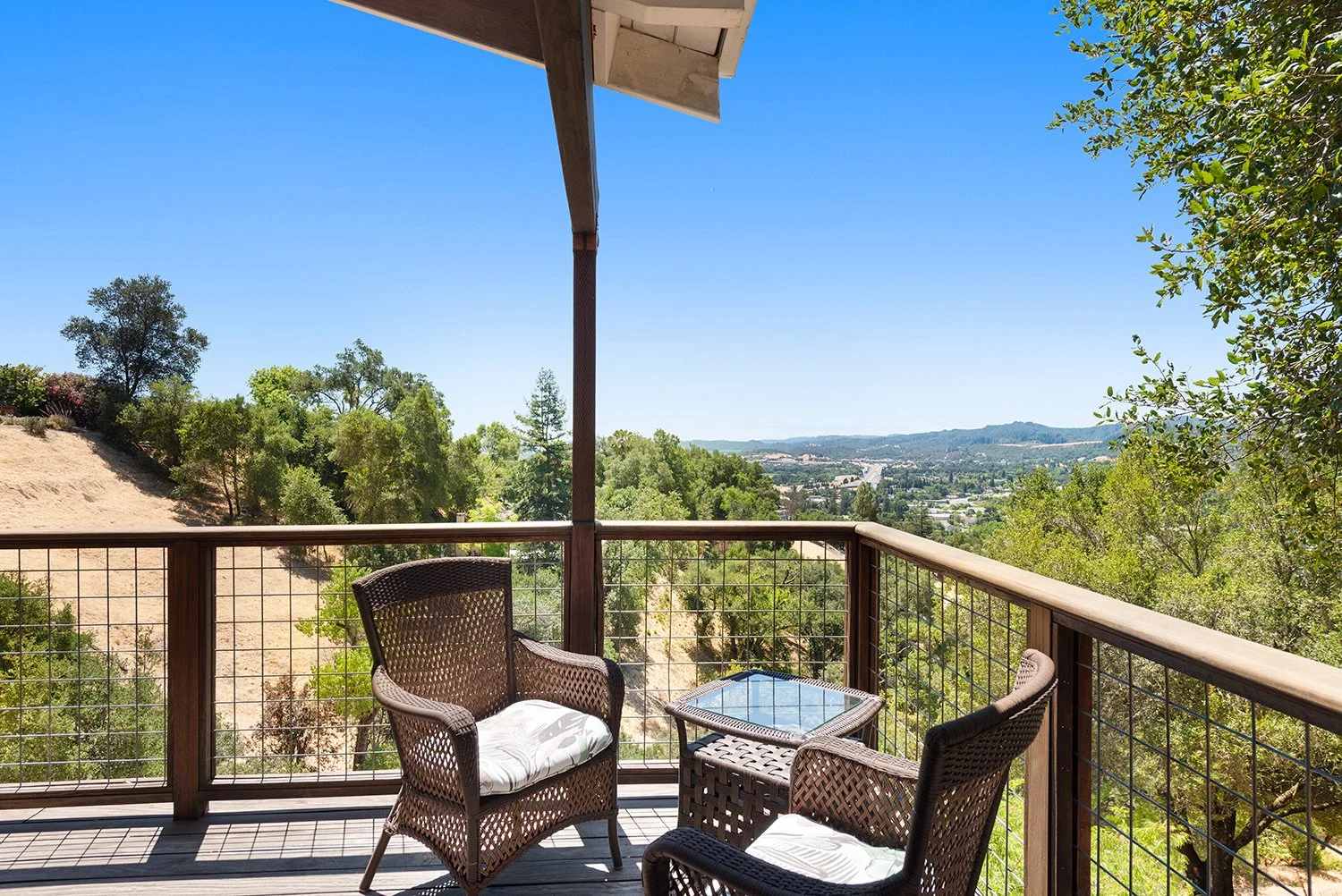 A balcony with wicker chairs and a glass-topped table, overlooking a scenic view of trees, hills, and a clear blue sky.
